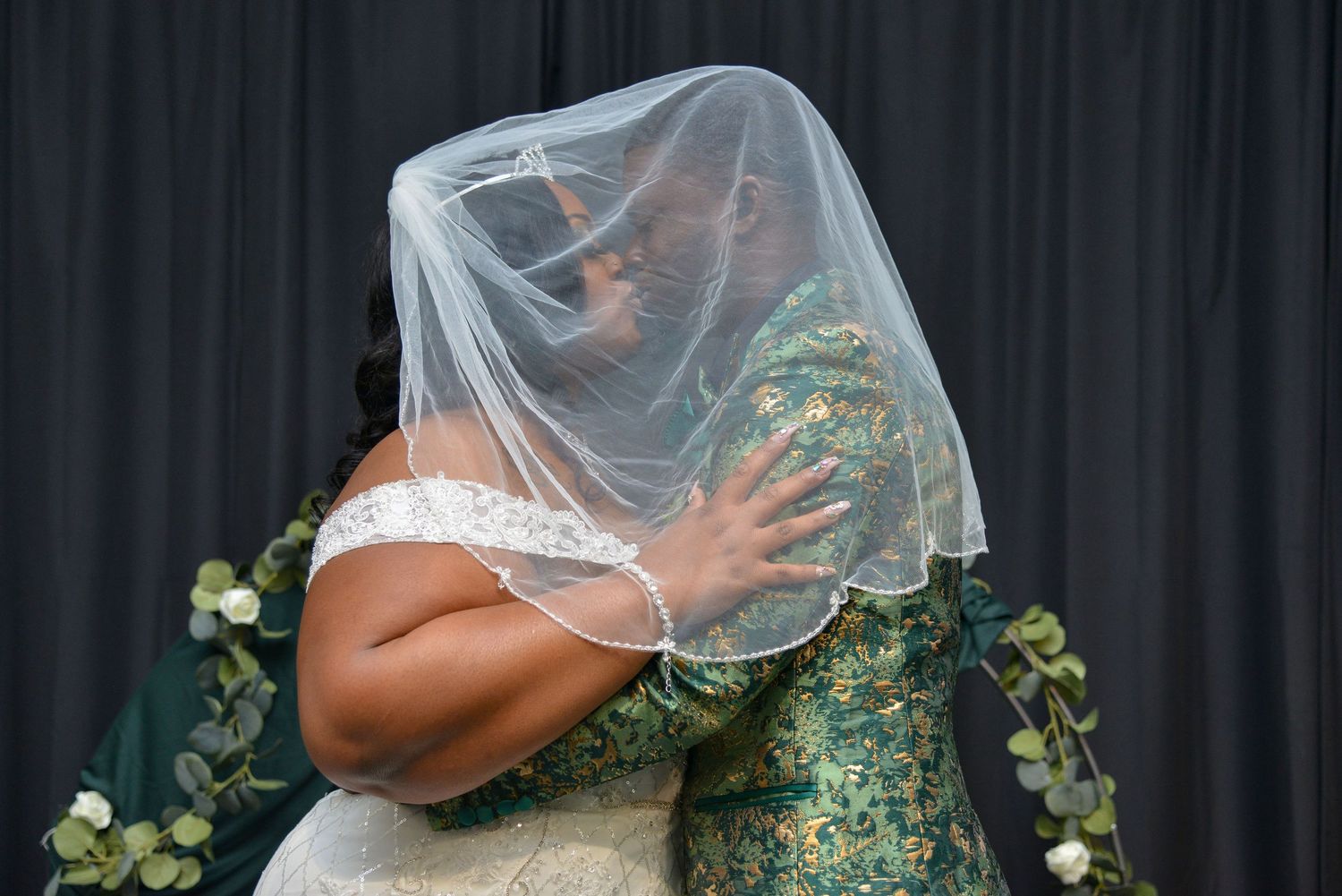 Groom and  bride share an emotional embrace under a wedding veil during their ceremony, one in white gown and one in green lace.