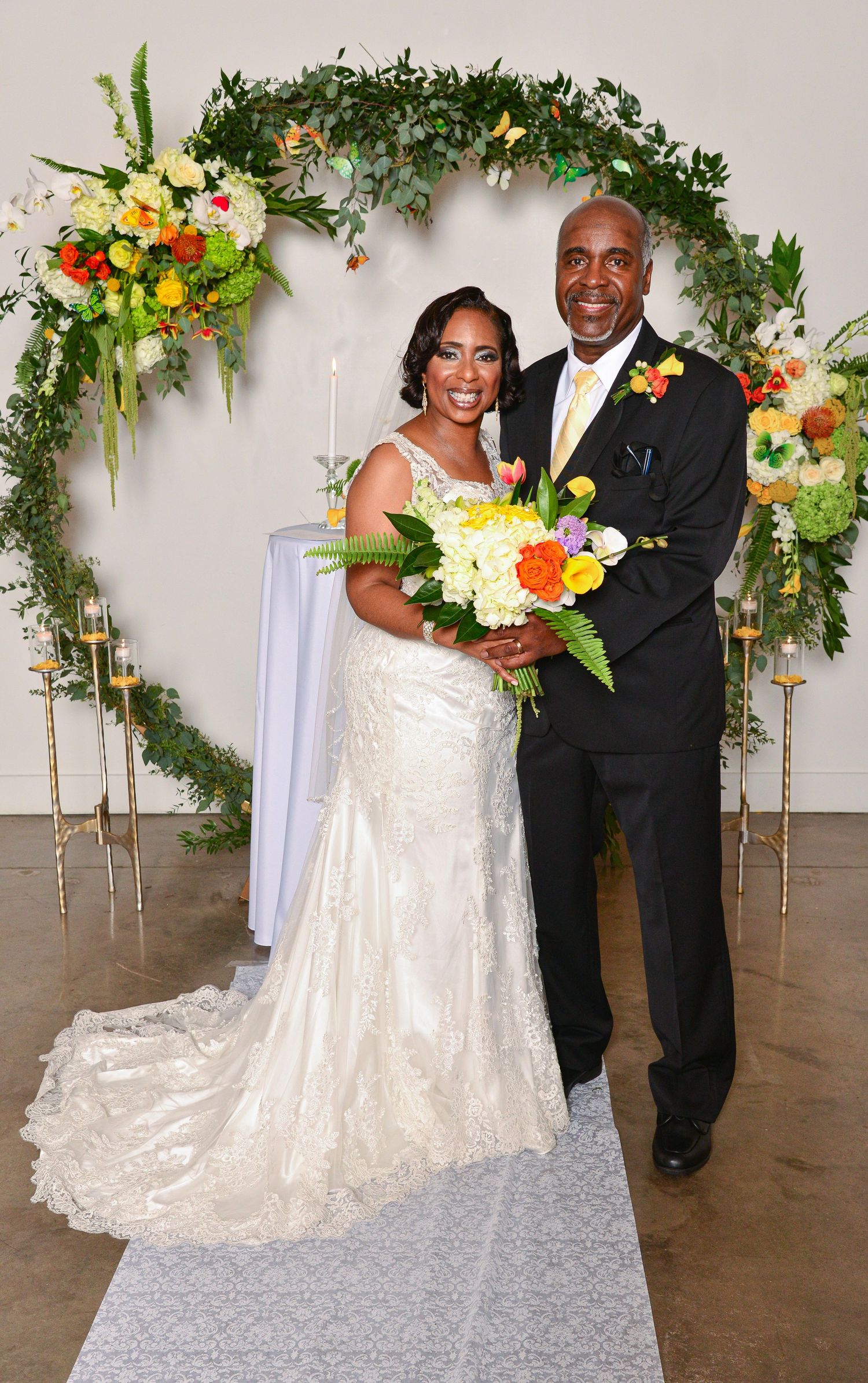 Bride and groom posing at wedding altar with colorful floral arch and bouquet.