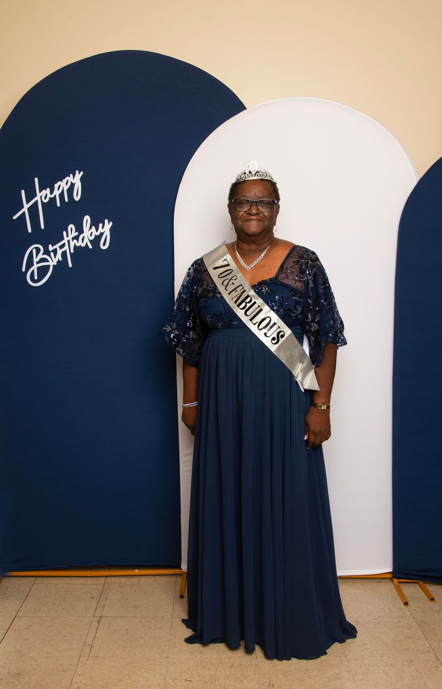 Woman in navy sequin gown and tiara wearing a birthday sash poses in front of Happy Birthday backdrop.