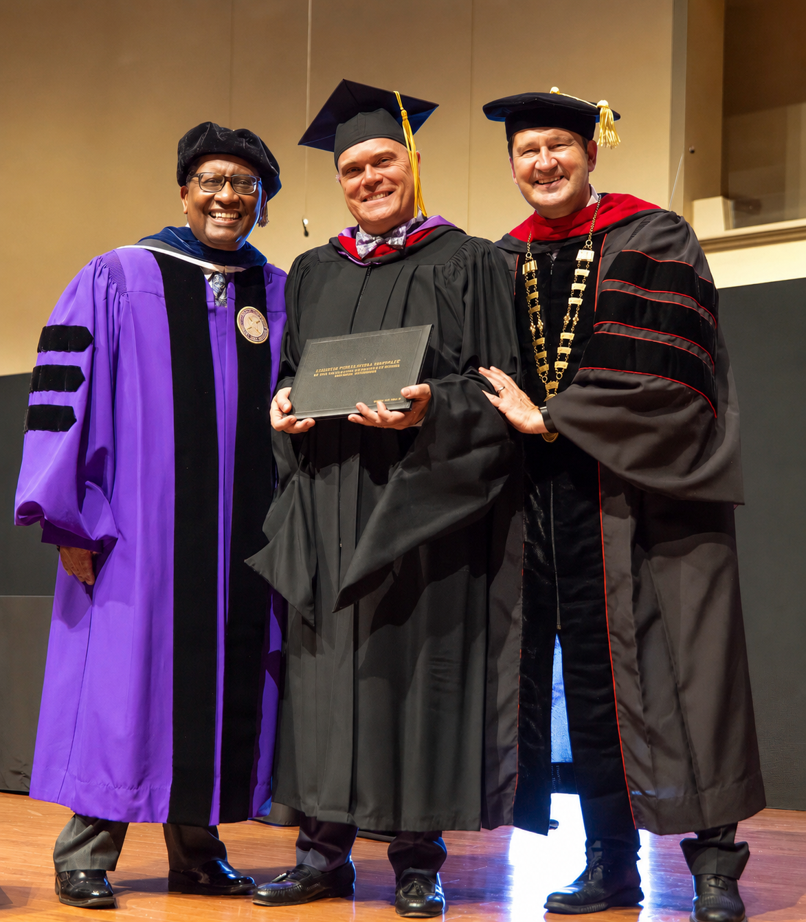 Three people in academic regalia smile at graduation ceremony, center graduate holds diploma flanked by two professors.