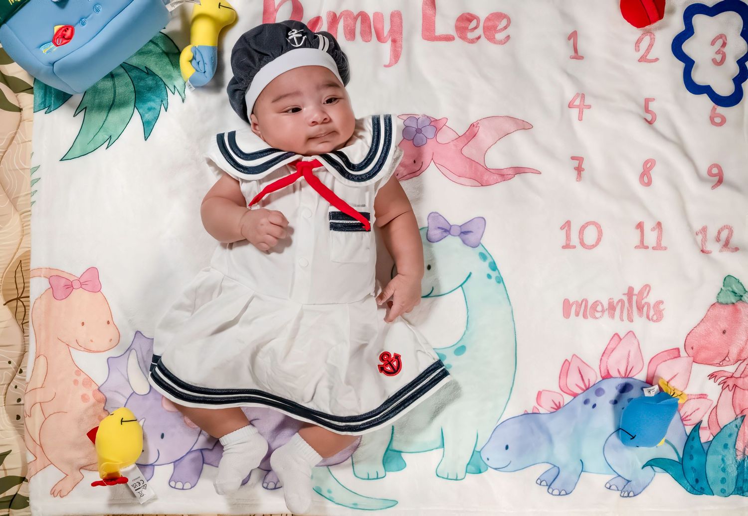 Baby in a sailor dress lying on a dinosaur milestone blanket for a monthly photo session.