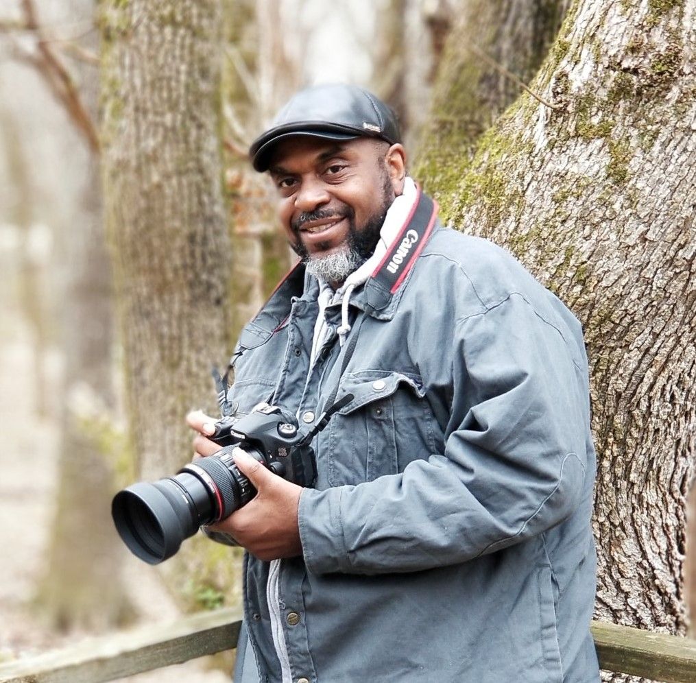 Male photographer smiling outdoors holding Canon camera near trees.