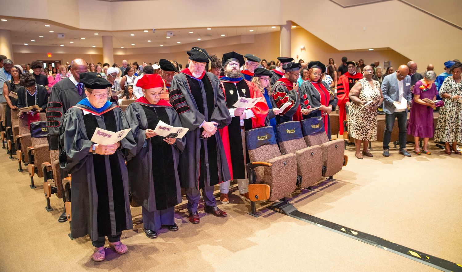 Faculty in colorful academic regalia standing in church aisle during graduation ceremony.