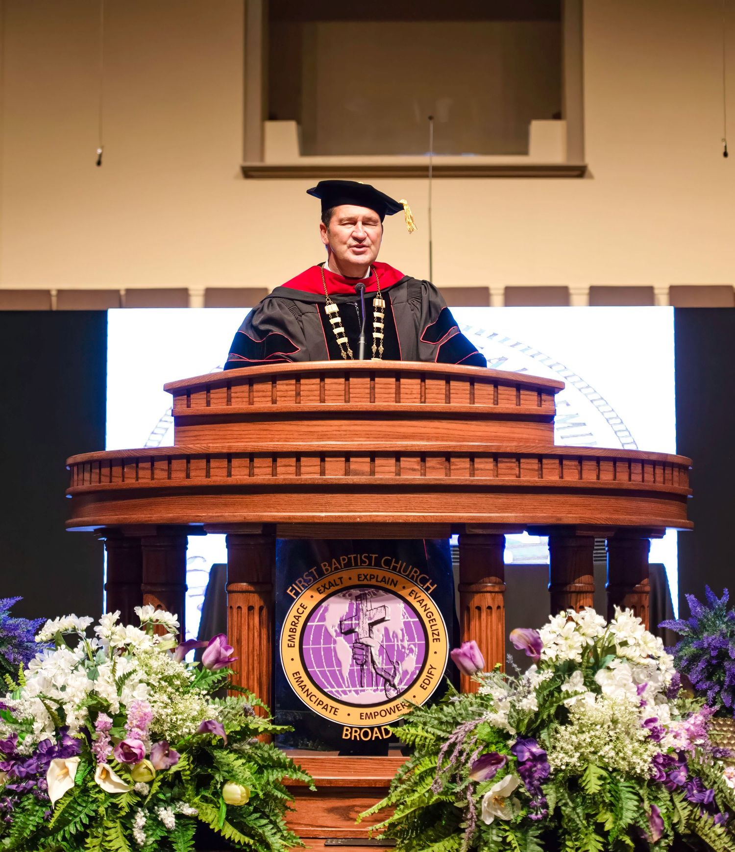Man in academic regalia speaks at ornate wooden podium during college graduation ceremony with floral arrangements.