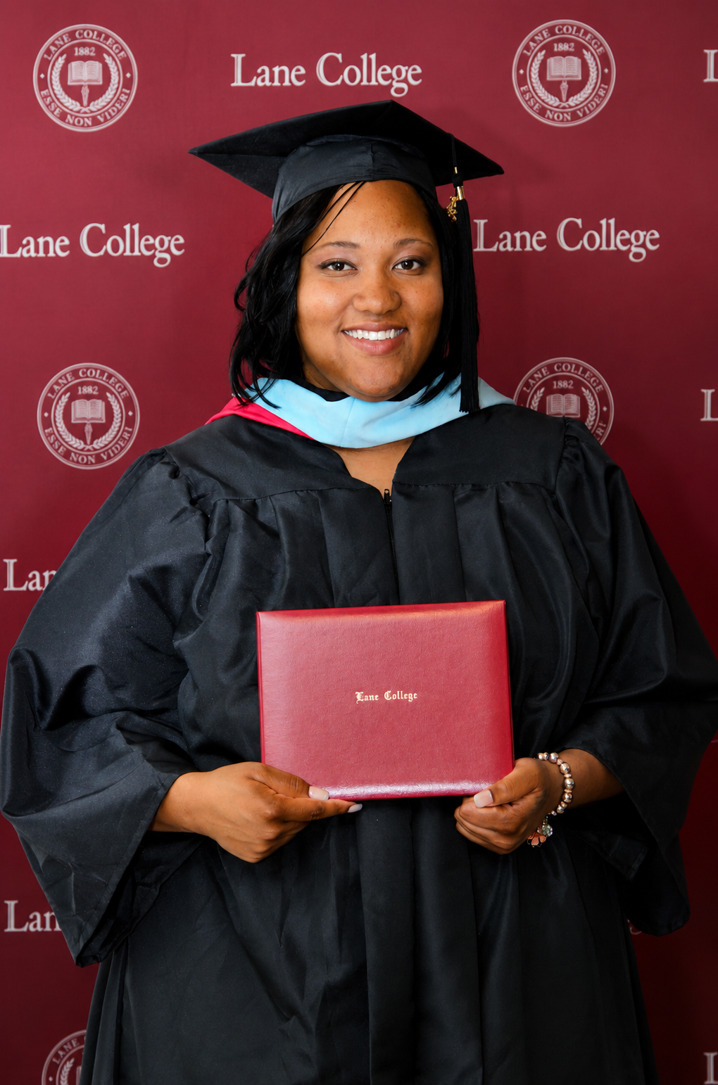 Smiling woman in black graduation gown holding Lane College diploma in front of Lane College banner.