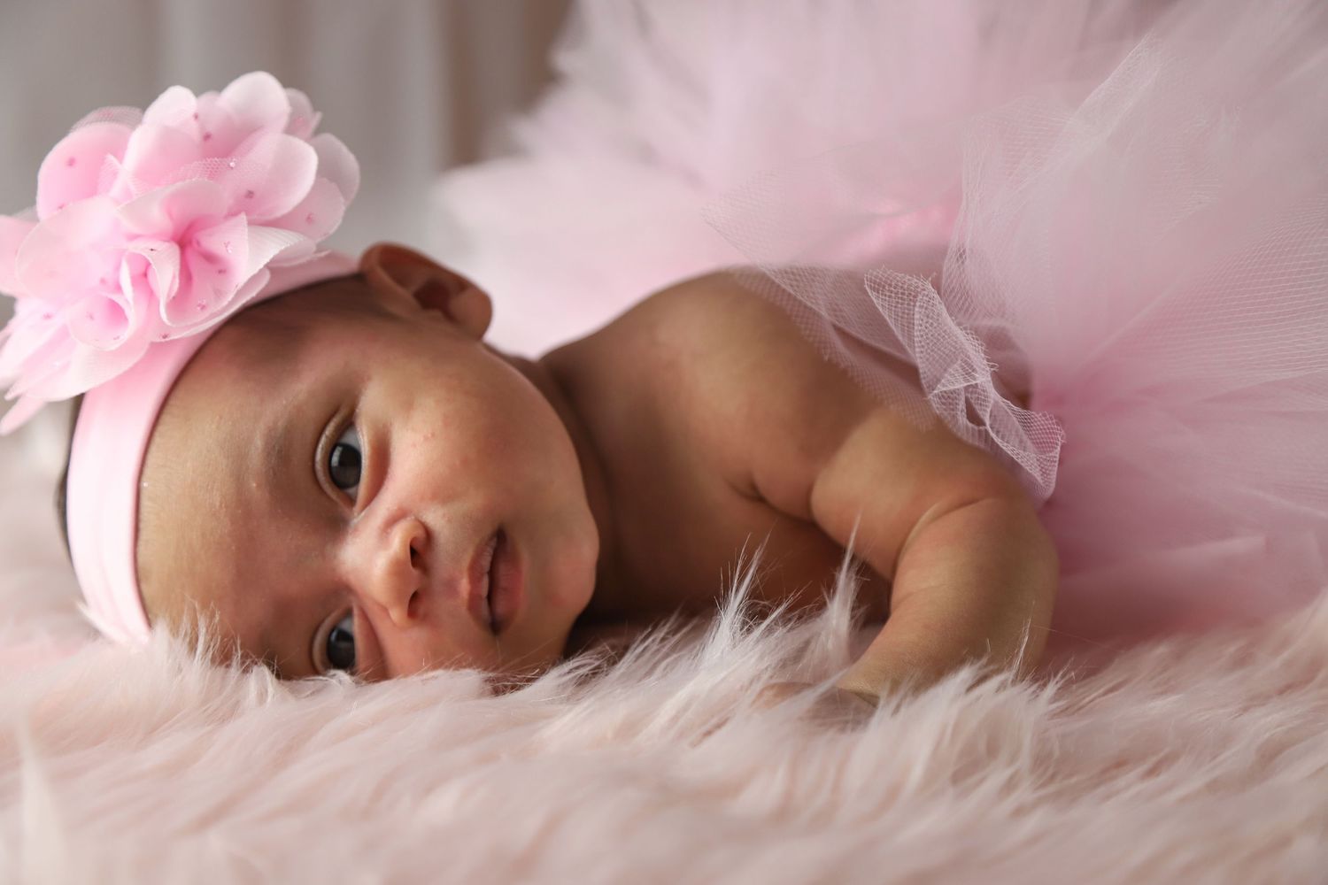 Adorable newborn baby girl in pink tutu skirt and floral headband lying on fluffy white fur blanket.