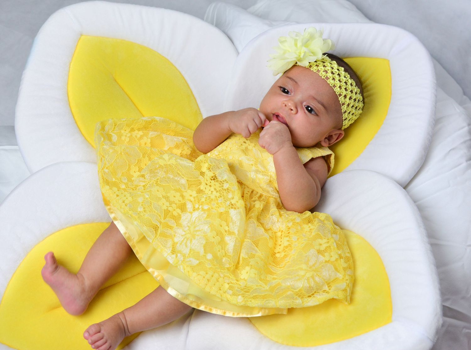 Cute baby in a yellow lace dress with floral headband lying on a flower-shaped white cushion.