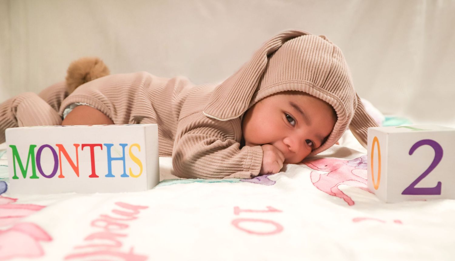 Baby in a beige bunny hoodie lying next to month milestone blocks on a decorative blanket.