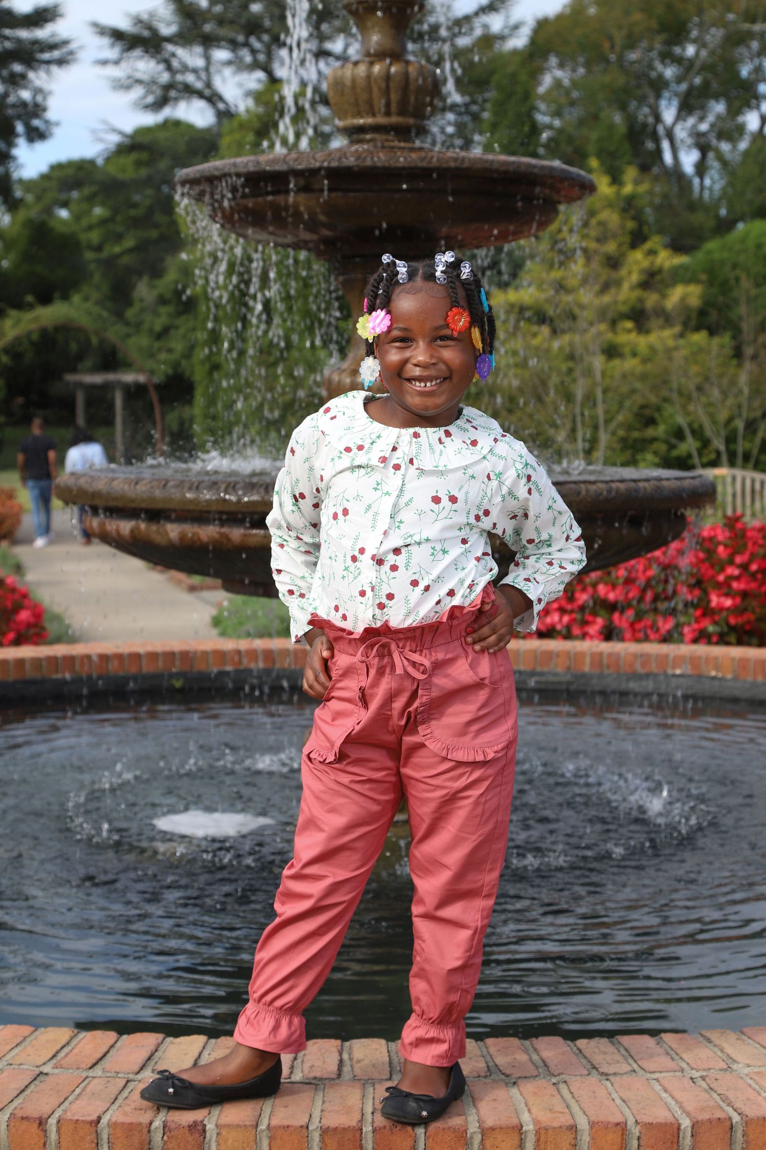 Happy young girl posing confidently in front of a garden fountain in pink pants.