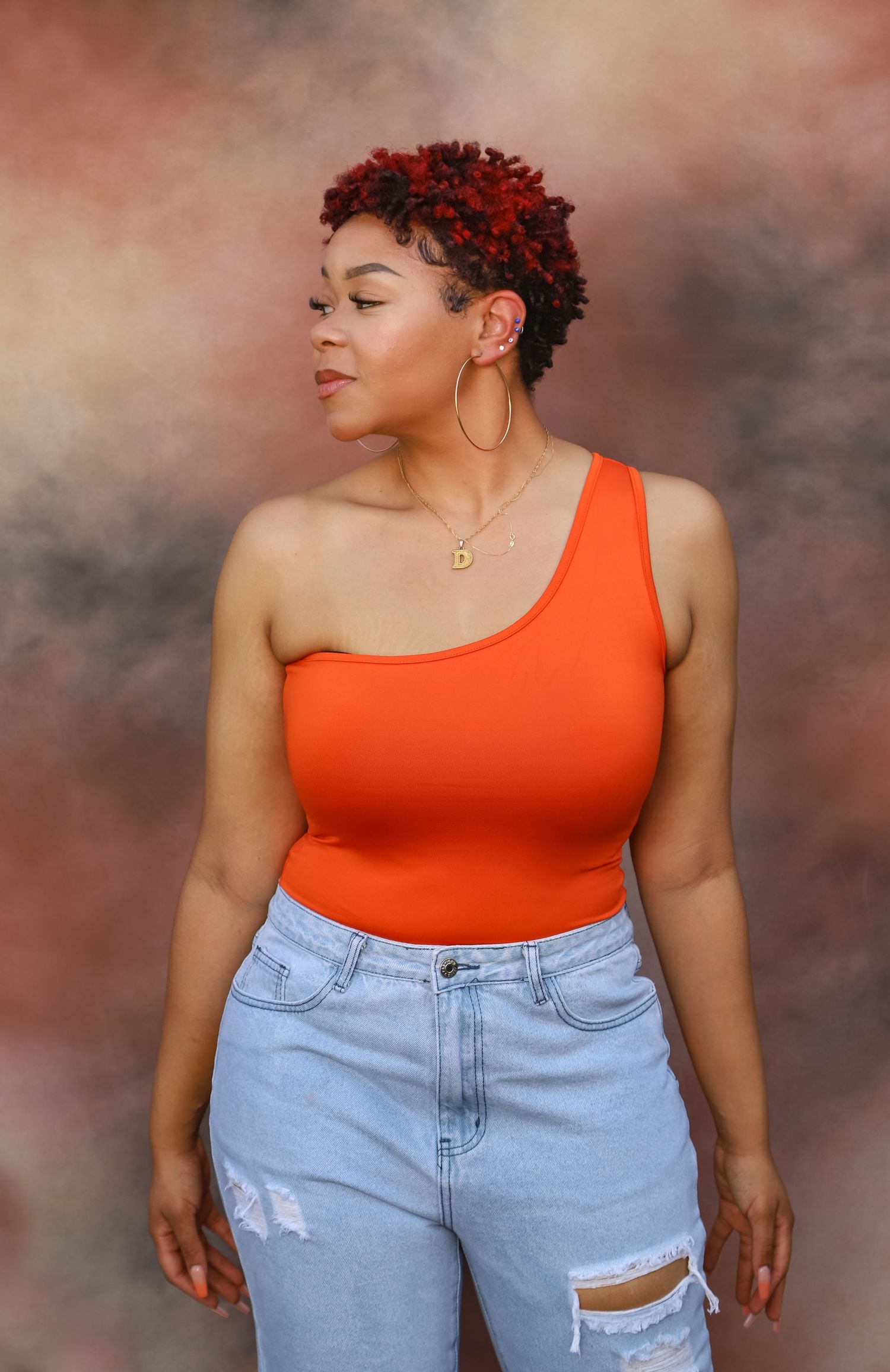 Woman in orange one-shoulder bodysuit and ripped jeans posing against colorful backdrop.