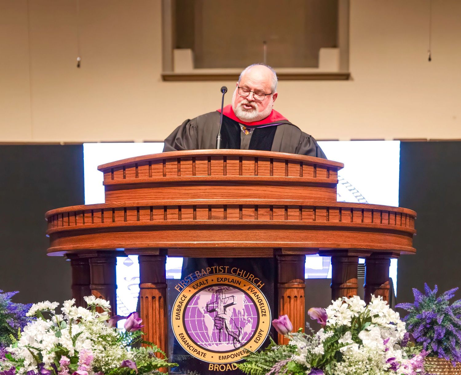 Male pastor in academic robes delivering sermon at ornate Baptist church pulpit.