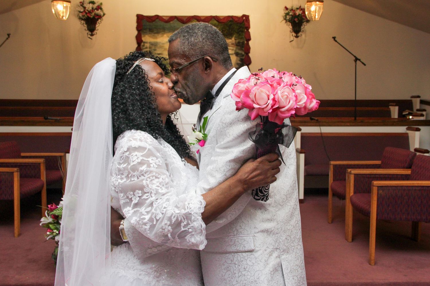 Couple sharing a romantic kiss in church holding pink rose bouquet at wedding.
