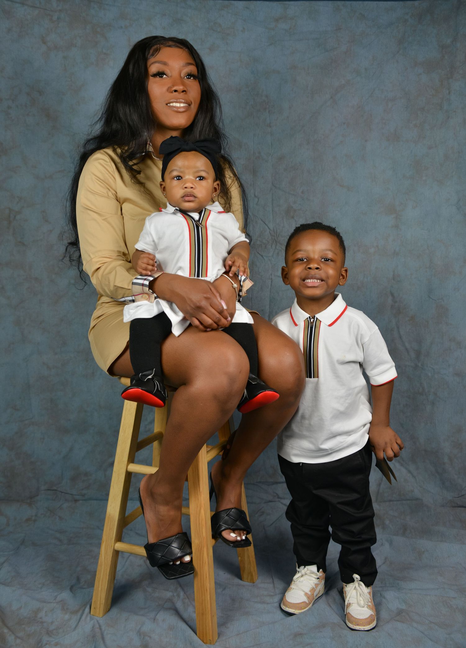 Mother sits on stool holding infant while toddler stands beside her in matching outfits.