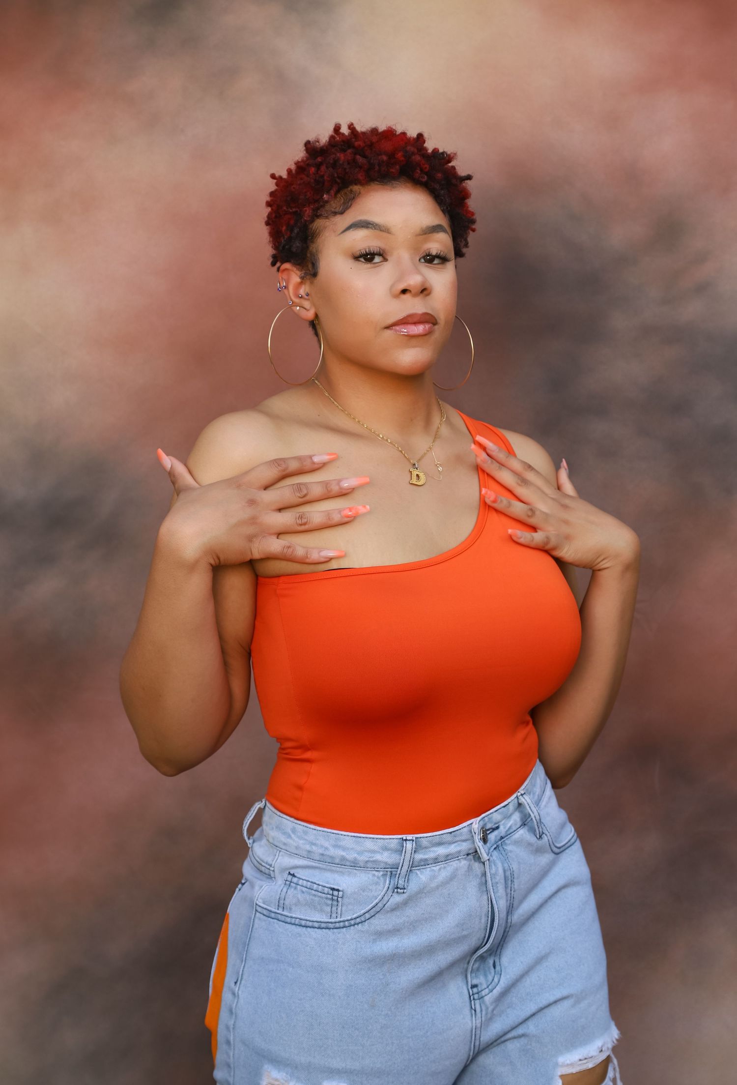 Young woman with red natural hair wearing orange bodysuit and denim shorts posing in studio.