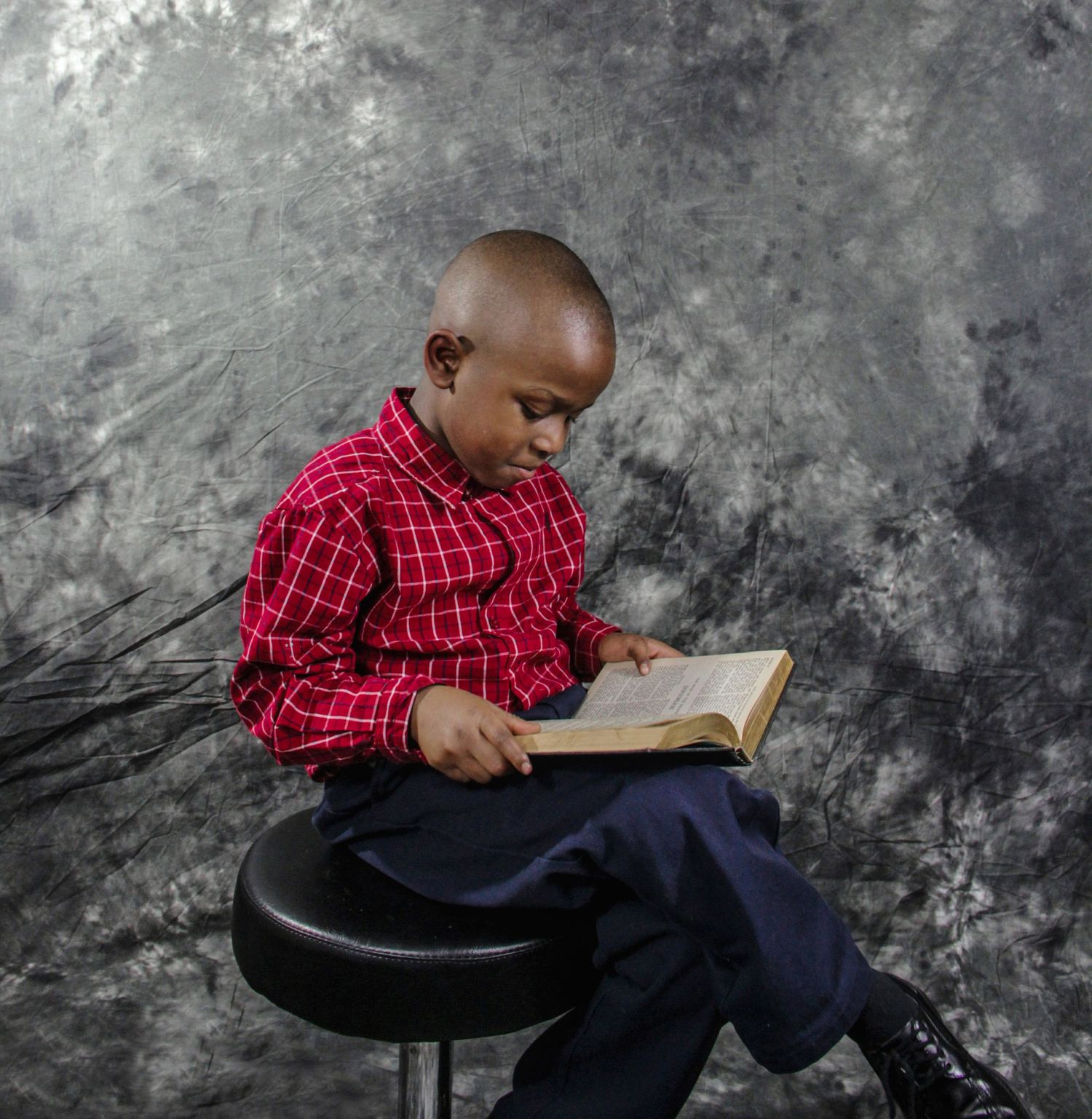 Young boy in red plaid shirt sits on stool reading a book against gray studio backdrop.