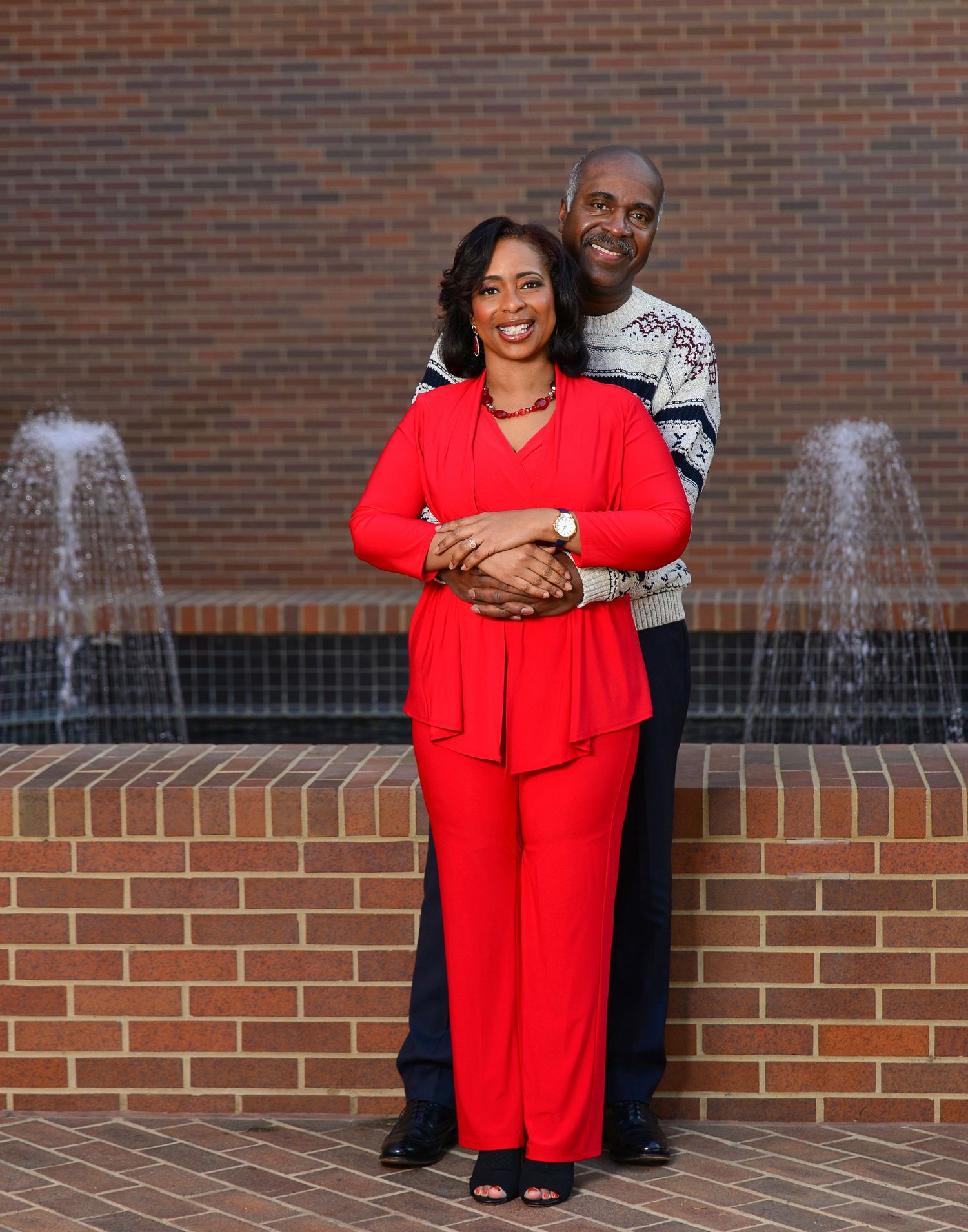 Couple posing romantically in front of brick wall fountains, woman in red pantsuit embraced by man in sweater.