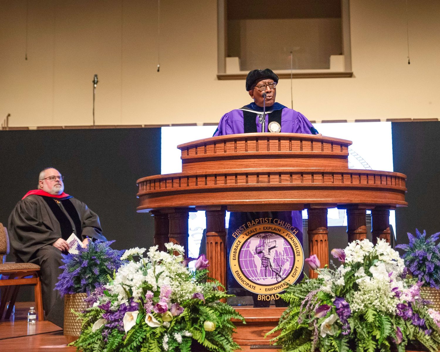Black male clergy in purple robes speaking at Baptist church pulpit during ceremony.