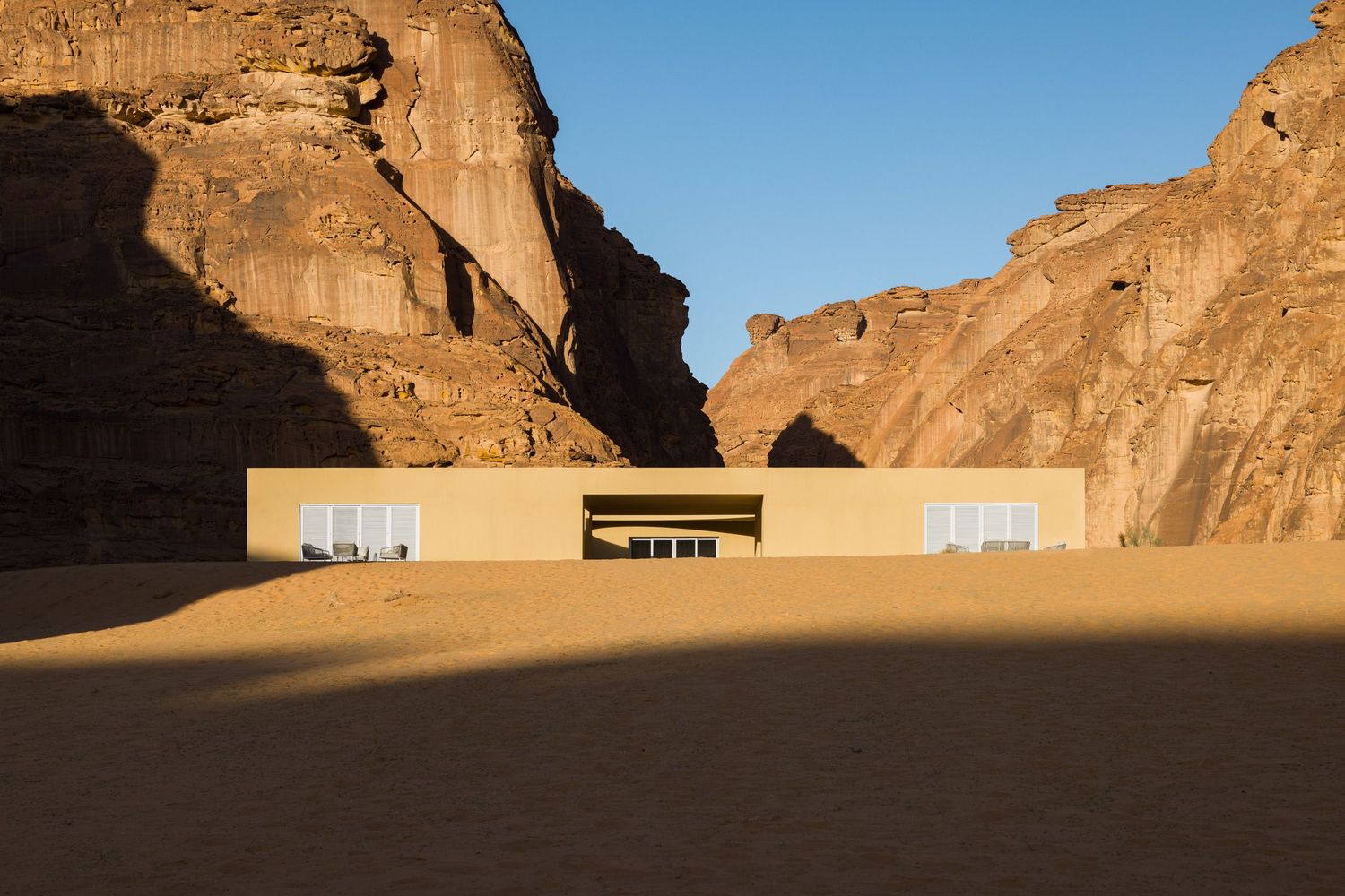 A sand-colored Desert X 2026 Visitor Center in AlUla Saudi Arabia emerging from a shadow during morning light, architecture by kwy.studio