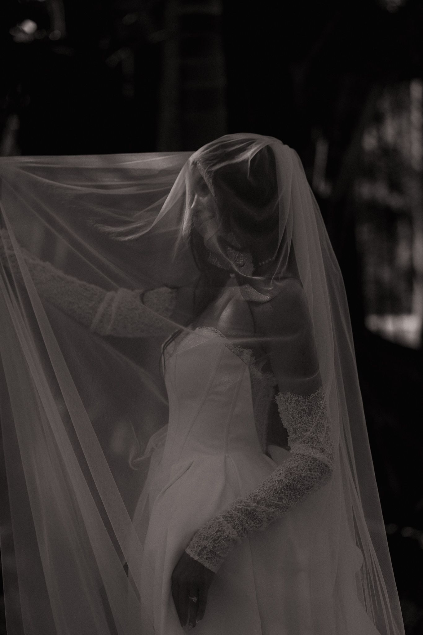 Black and white photo of bride in wedding dress with veil draped over couple embracing outdoors.
