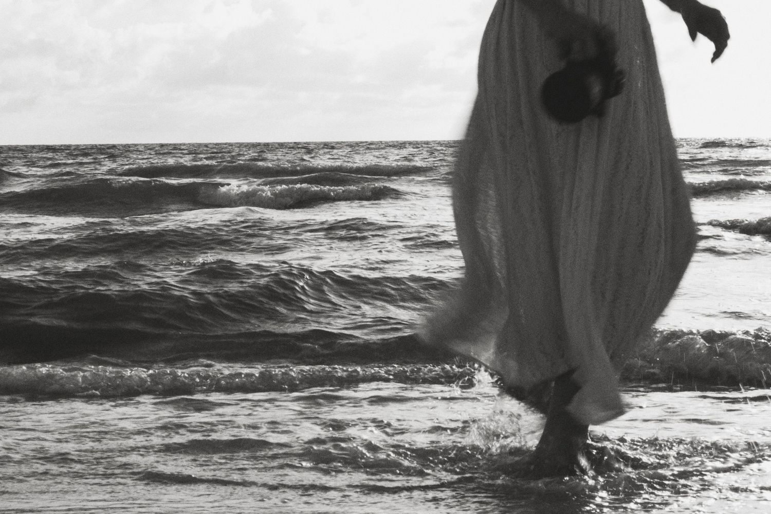 Black and white photo of a woman in a flowing white dress walking in ocean waves on a beach.