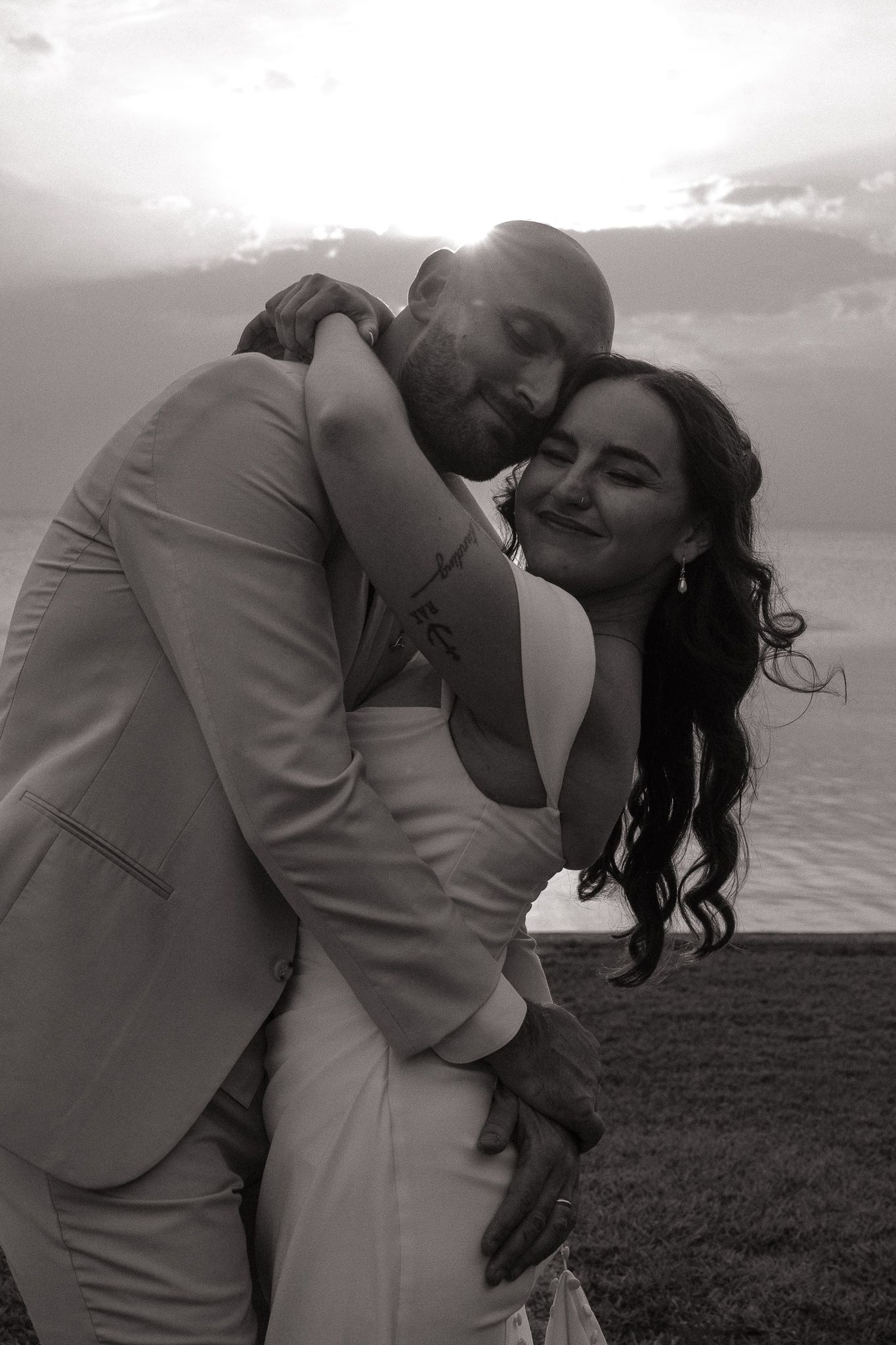 Black and white photo of a couple embracing at sunset near the ocean, dressed in wedding attire.