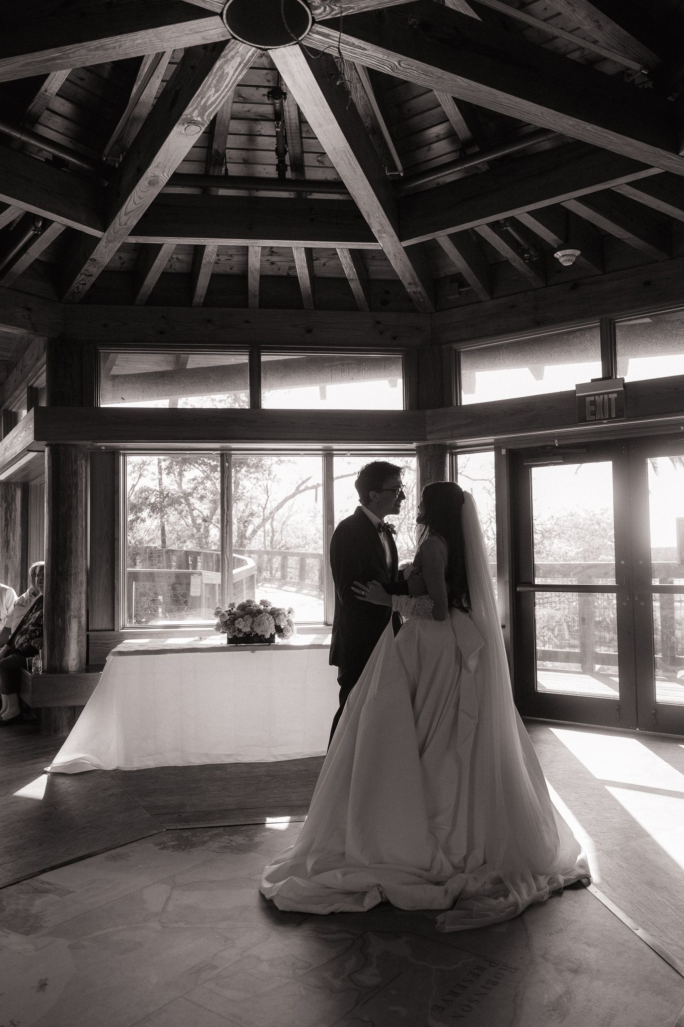 Bride and groom share an intimate embrace inside a rustic wooden pavilion with large windows in black and white.