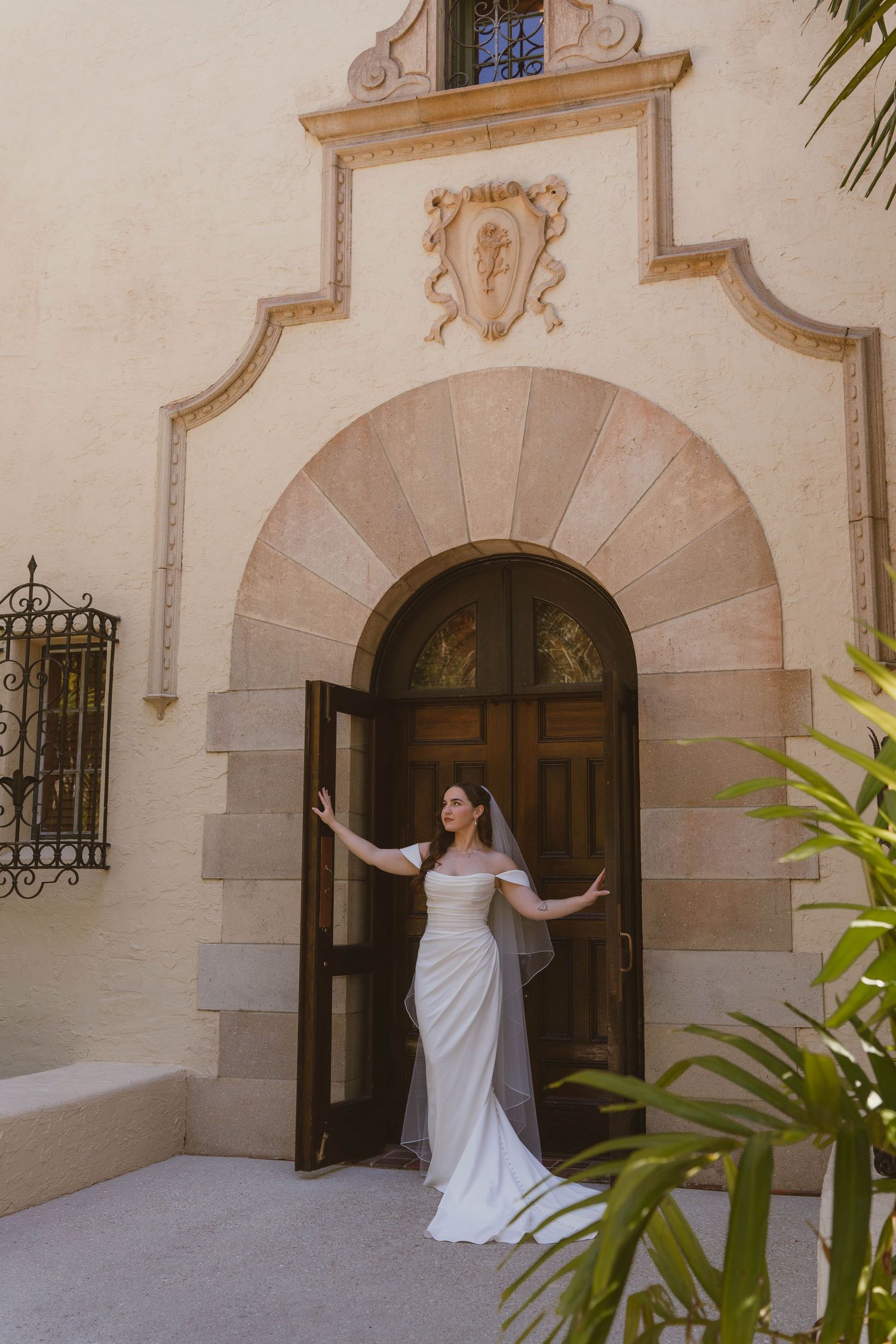 Radiant bride in white gown opening ornate wooden doors of elegant Mediterranean-style venue.