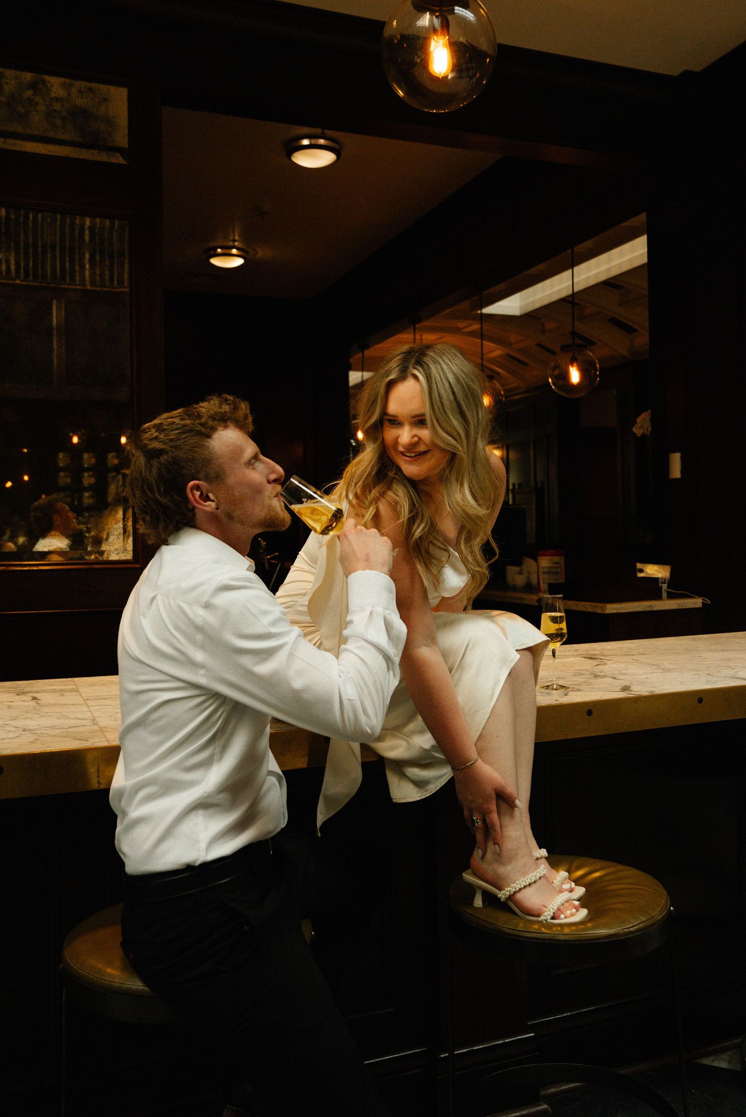 Romantic couple at a dimly lit bar, man kneeling beside woman seated on a barstool with champagne.
