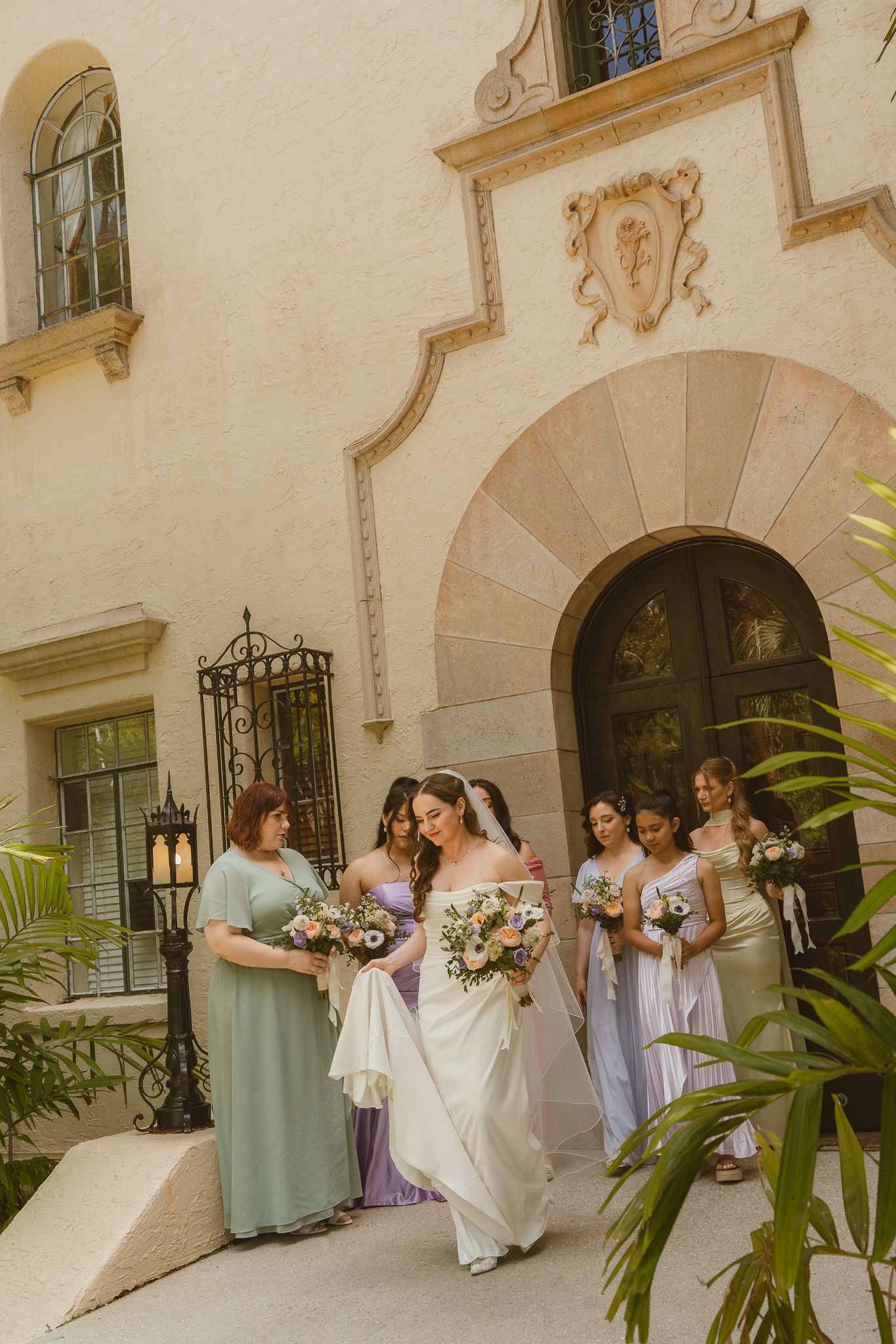 Bride in white gown walks with bridesmaids in lavender and sage dresses outside a Mediterranean venue.