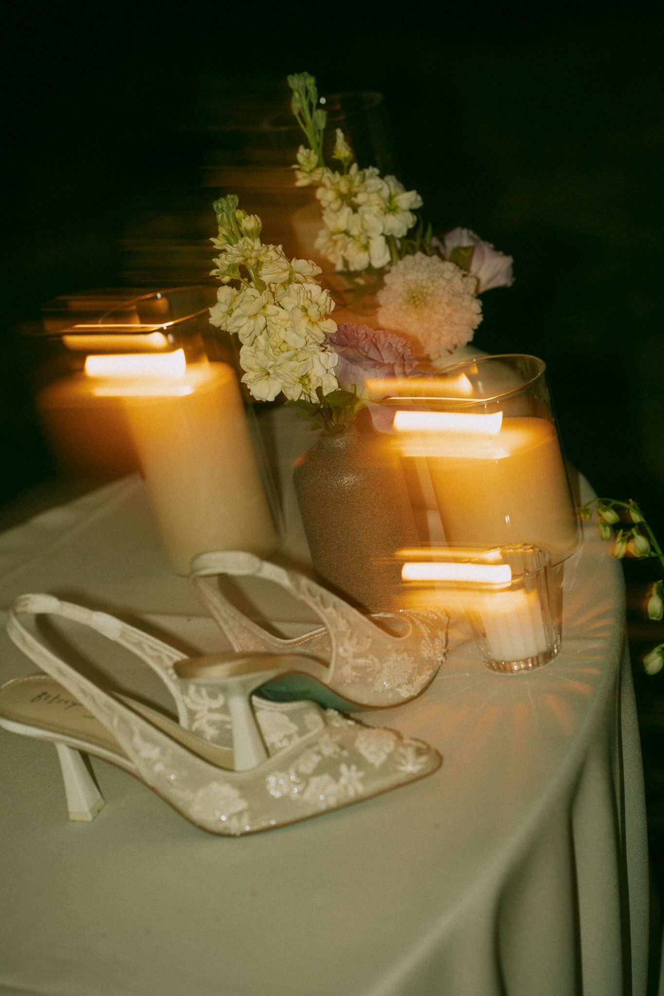 Elegant bridal heels with floral lace beside glowing candles and soft blooms on a white table.