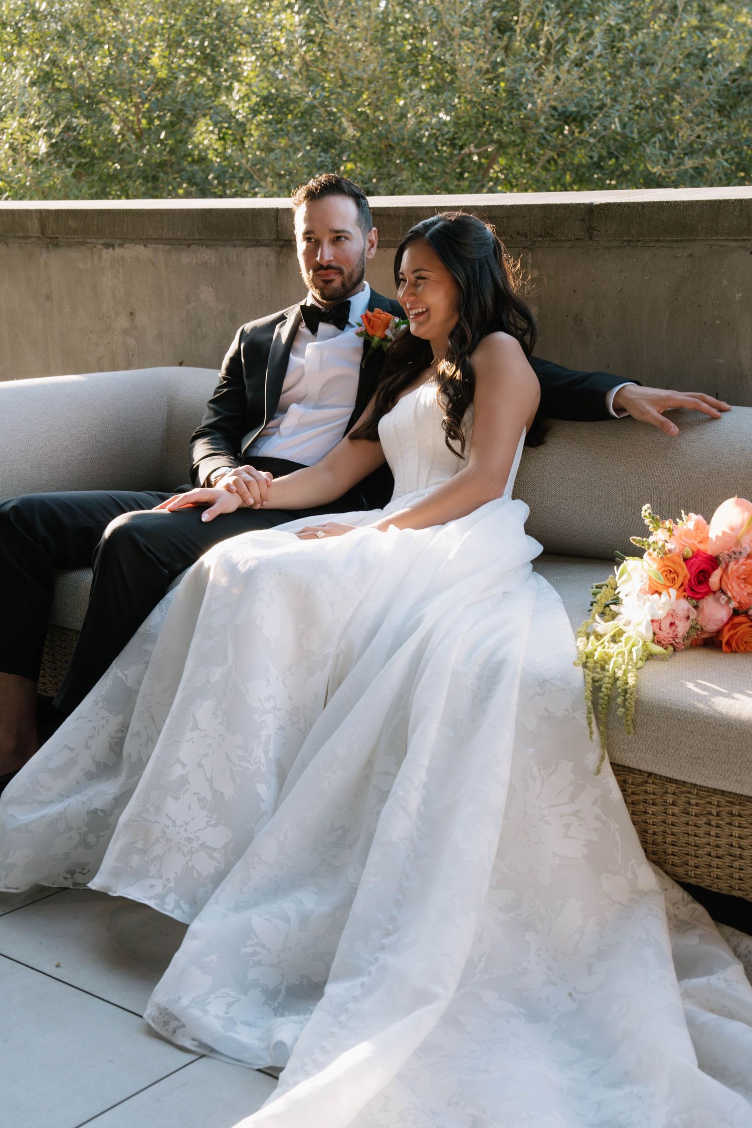 Bride and groom sit together on an outdoor terrace sofa, with a vibrant coral and pink bridal bouquet nearby.