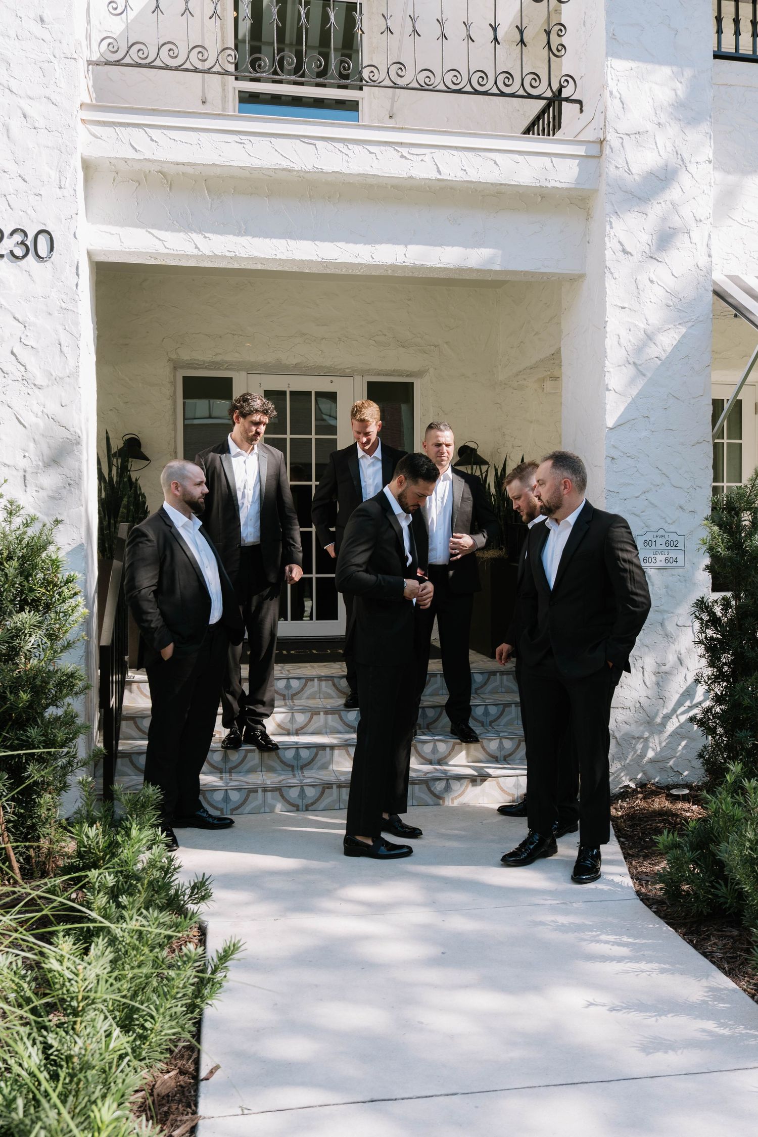 Groom and groomsmen in black suits gather outside a white building before the wedding ceremony.