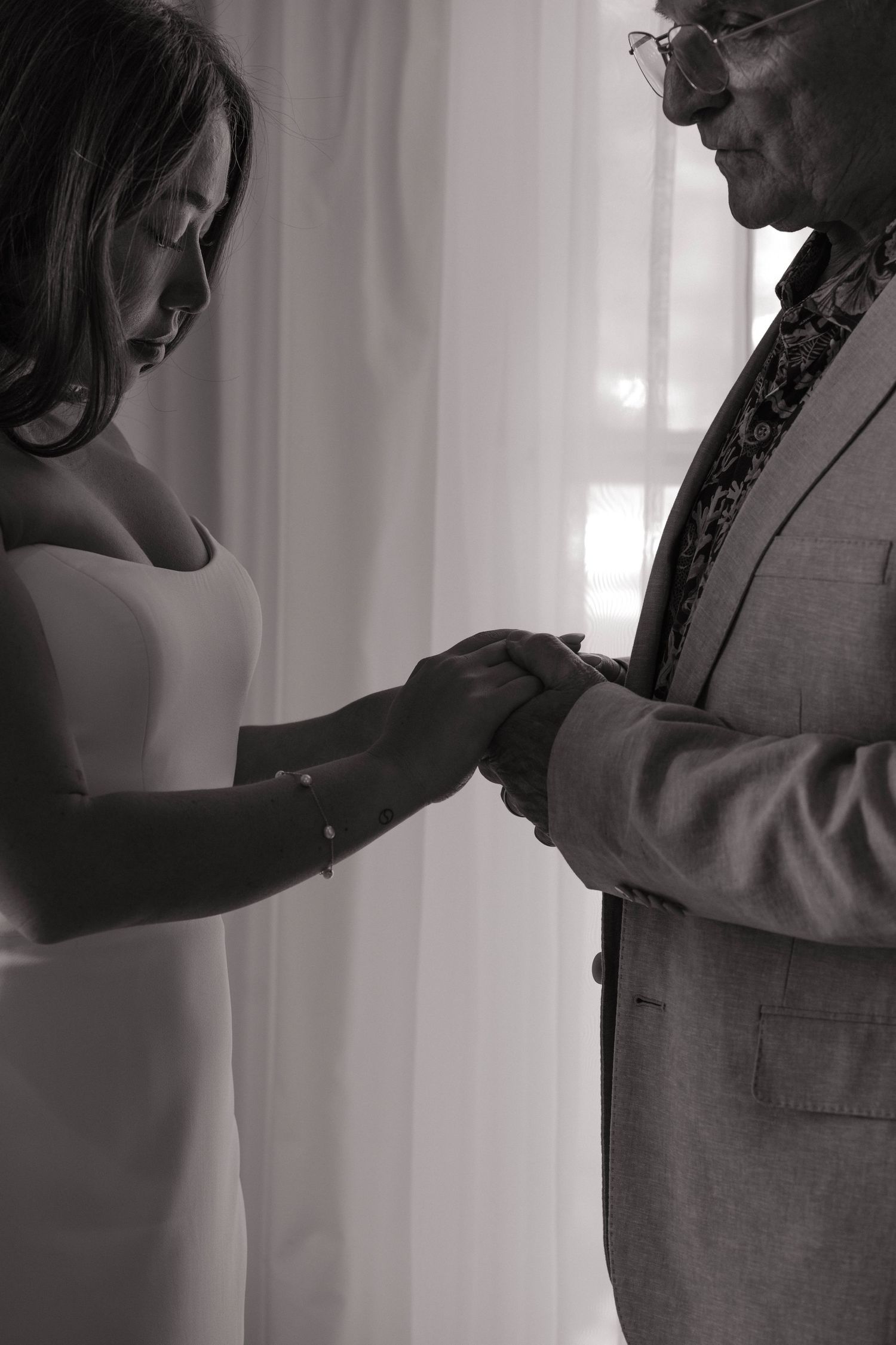 Bride and older man share tender hand-holding moment in black and white wedding photograph.