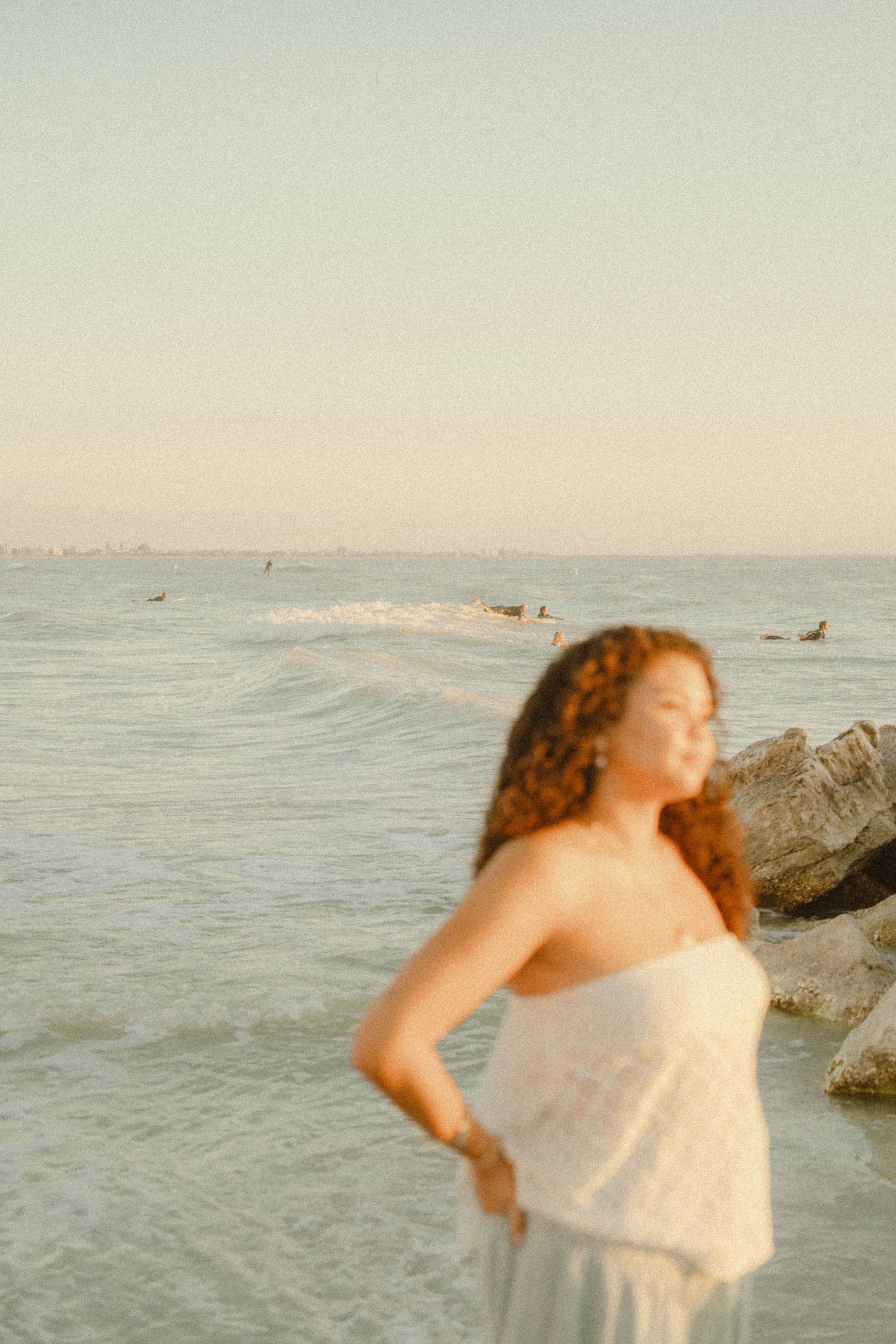 Blurred artistic shot of curly-haired woman in white dress standing by ocean rocks.