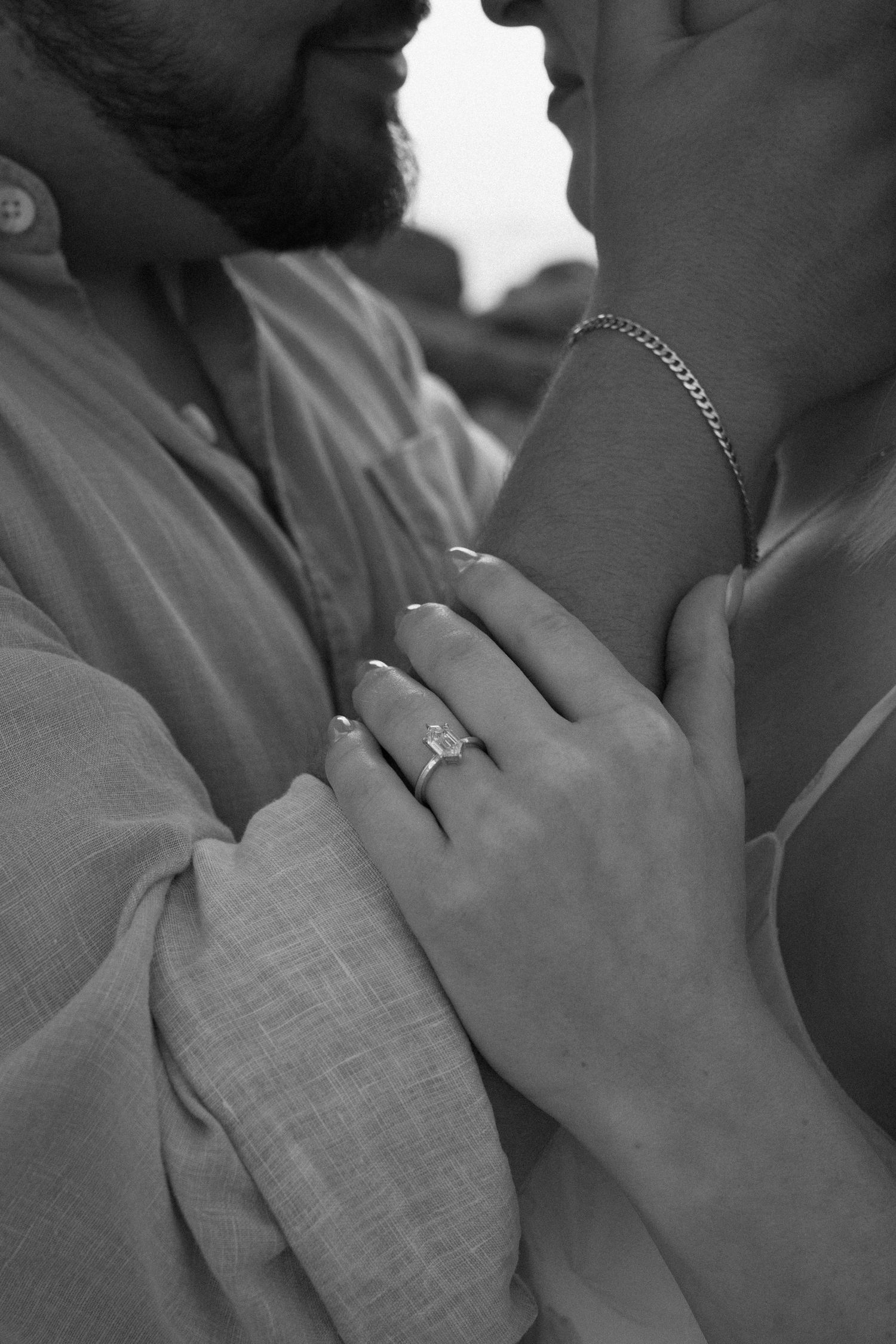 Black and white close-up of couple embracing with focus on engagement ring and gold chain bracelet.