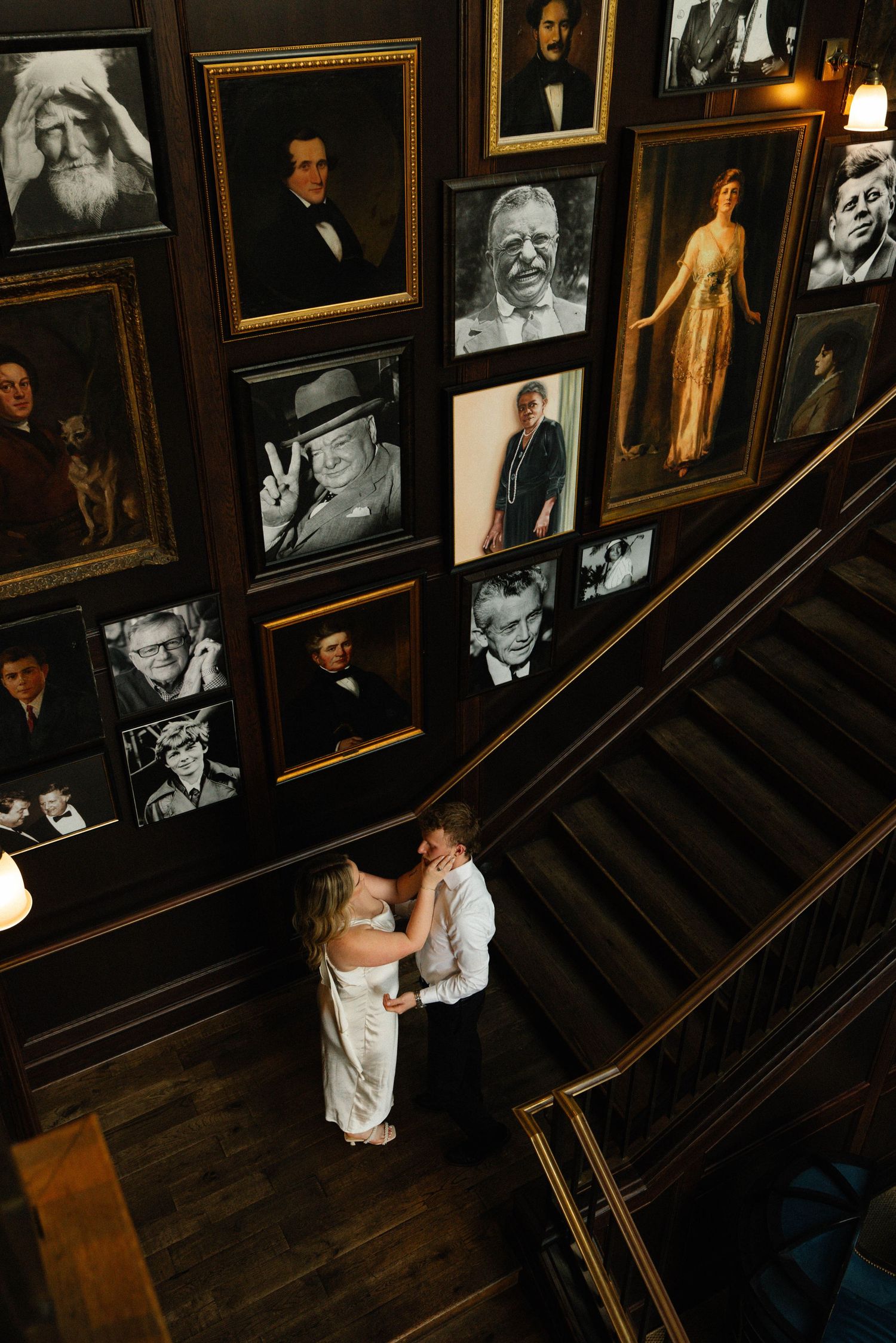 Bride and groom share a romantic moment on stairs surrounded by a gallery wall of framed portraits.