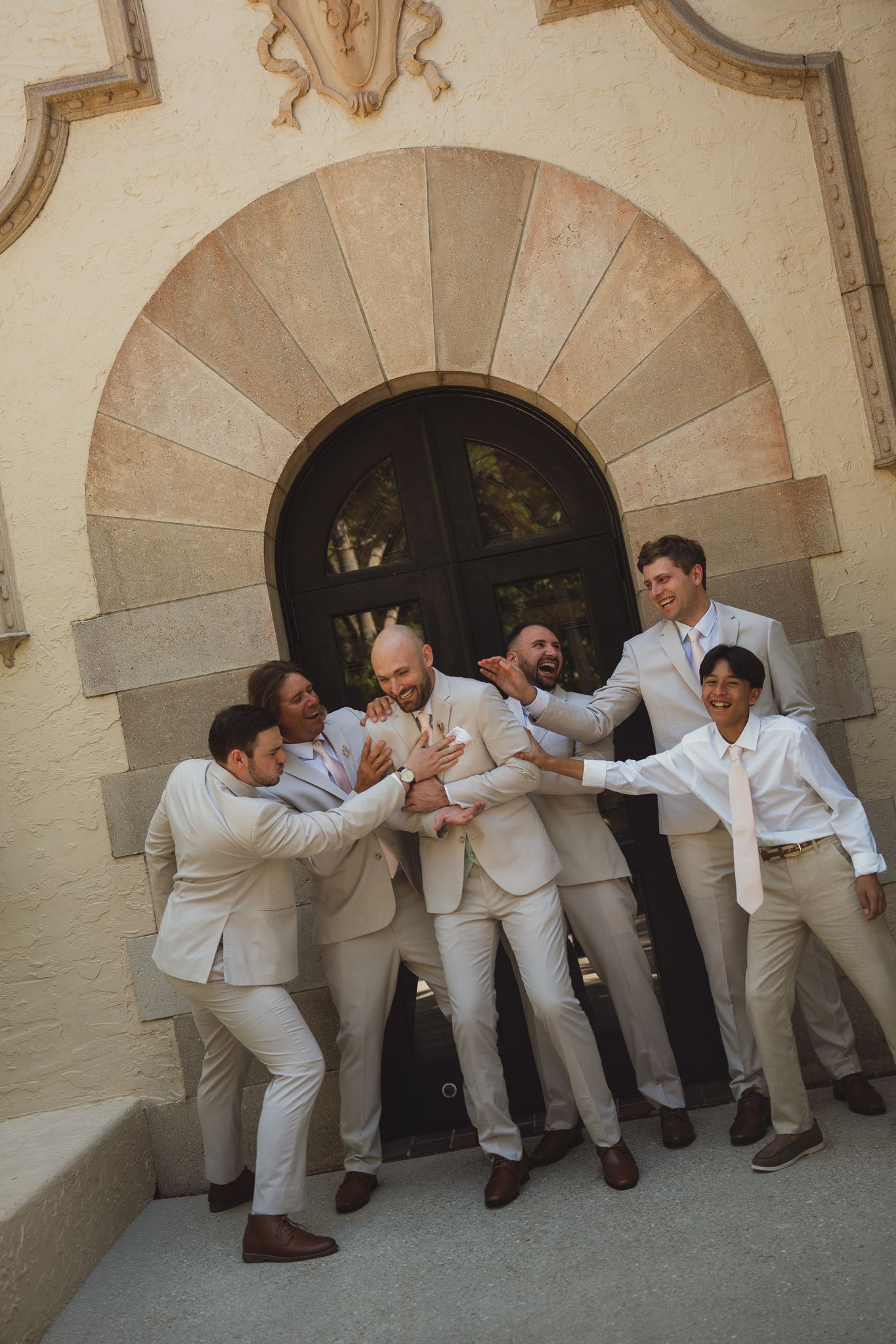 Groomsmen in beige suits playfully pose together in front of an arched doorway at a wedding venue.