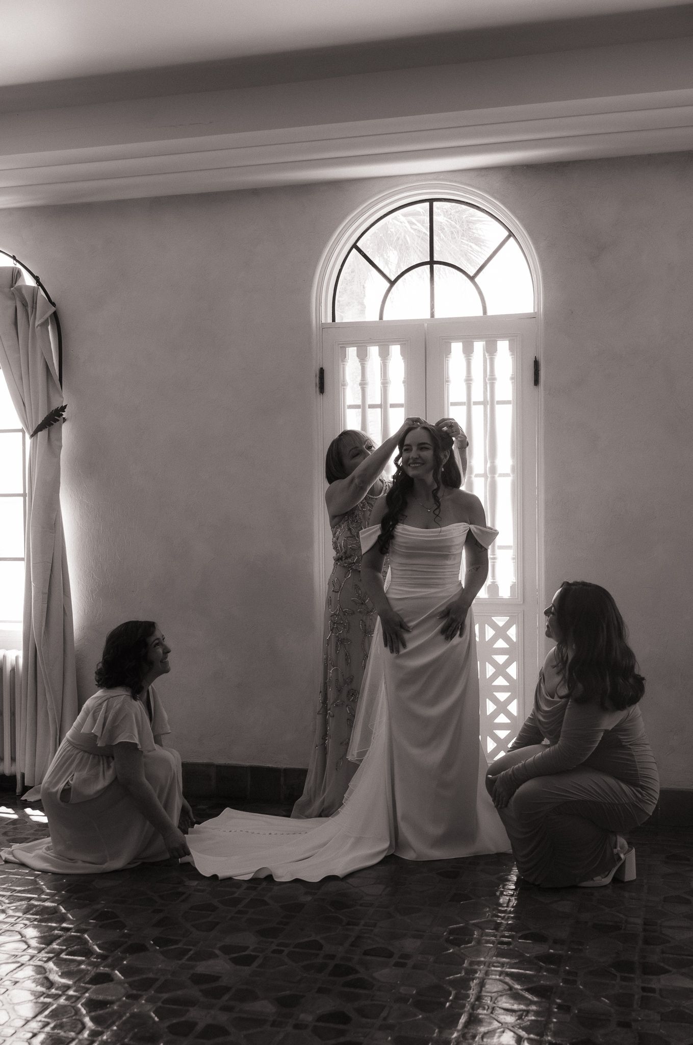 Black and white photo of a bride in a white gown being helped by bridesmaids near an arched window.