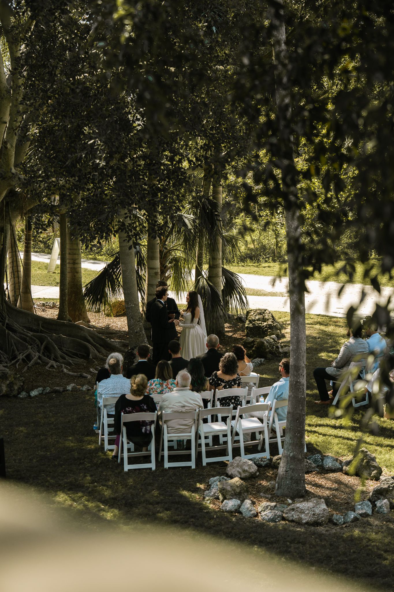 Intimate outdoor wedding ceremony with small guest gathering seated on white chairs beneath towering trees.