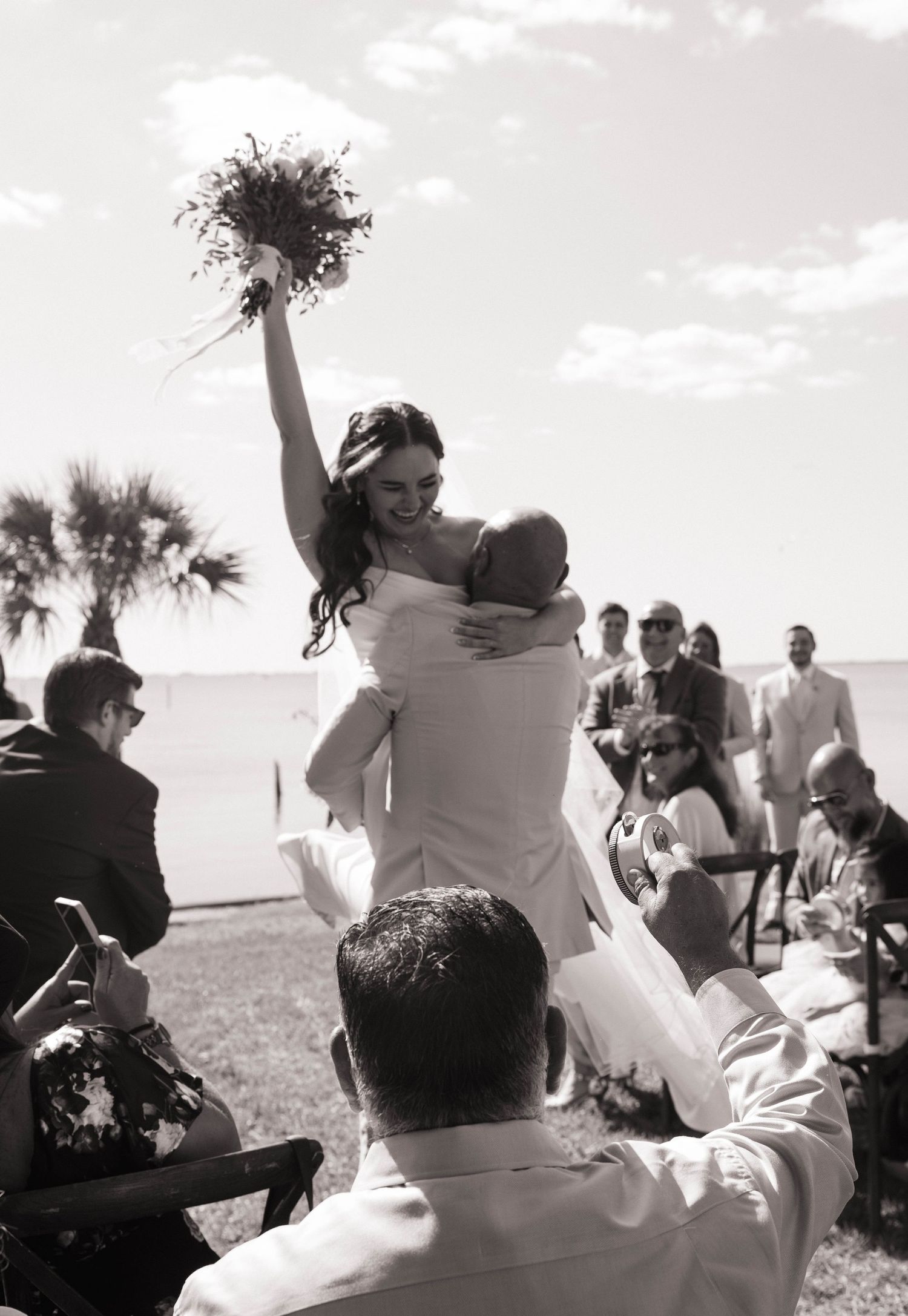 Black and white photo of jubilant bride lifted by groom raising bouquet at outdoor beach wedding ceremony.