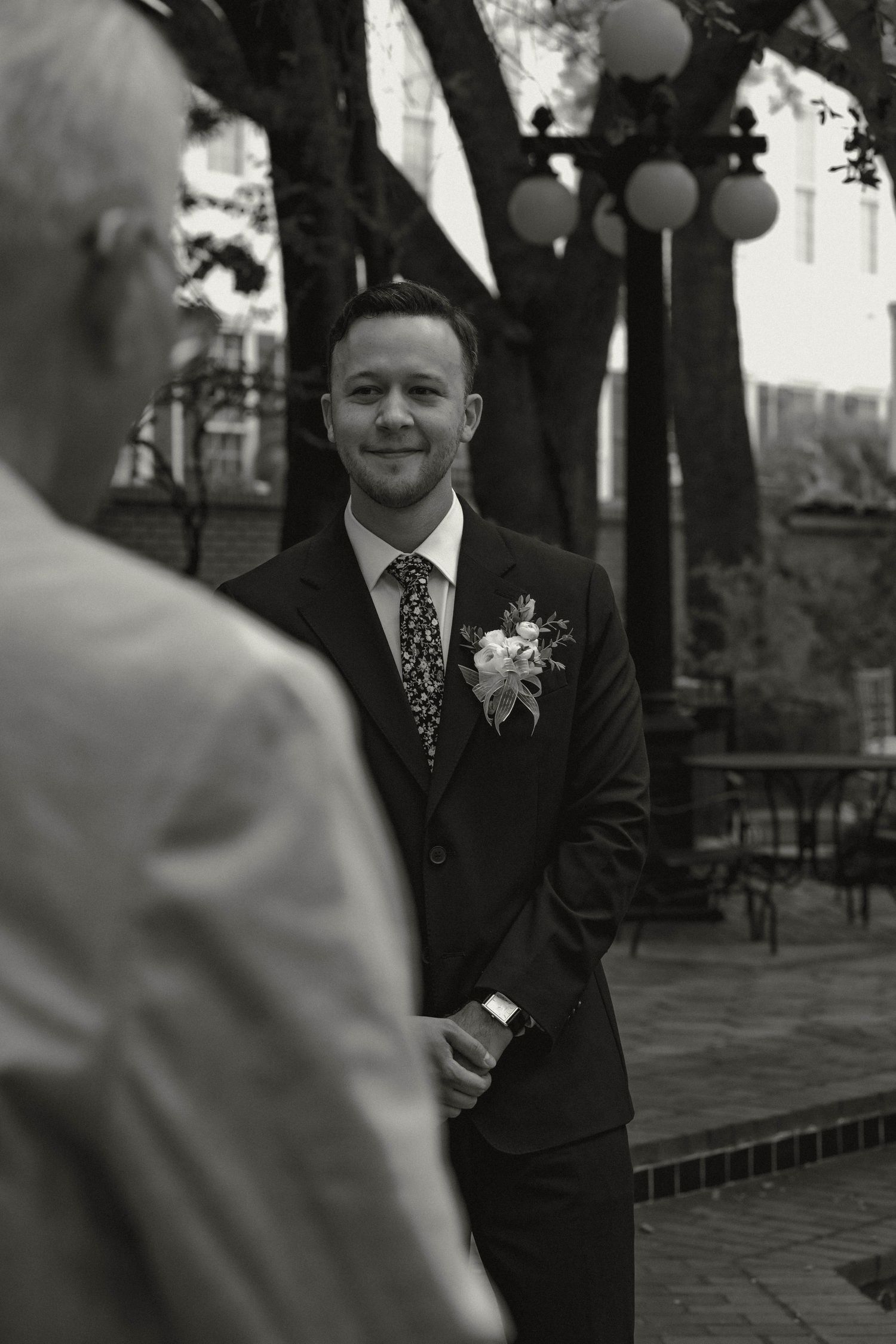 Smiling groom in dark suit with floral boutonniere stands outdoors during black and white wedding ceremony.