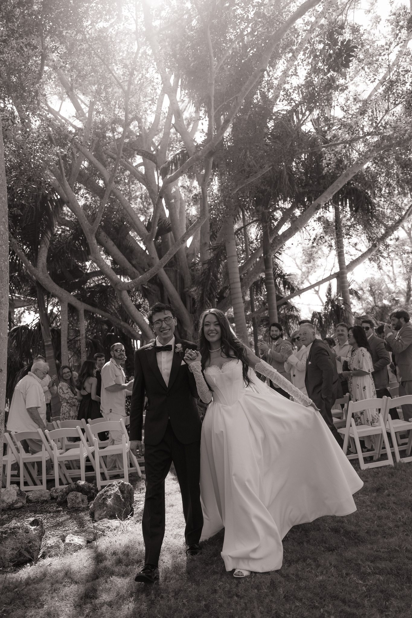 Black and white photo of newlyweds walking down outdoor aisle surrounded by guests and trees.
