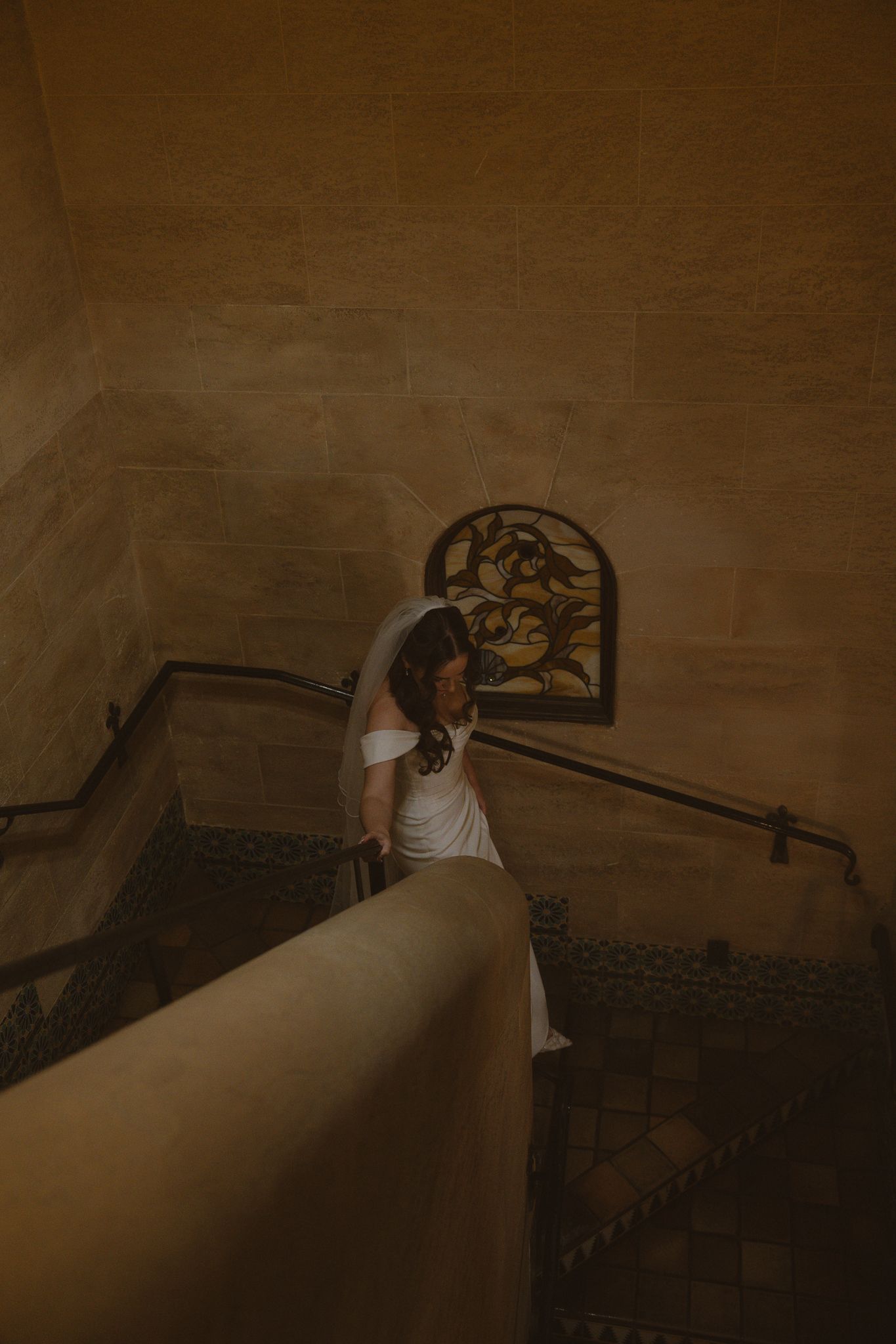Bride in white gown and veil ascending ornate stone staircase in elegant venue.
