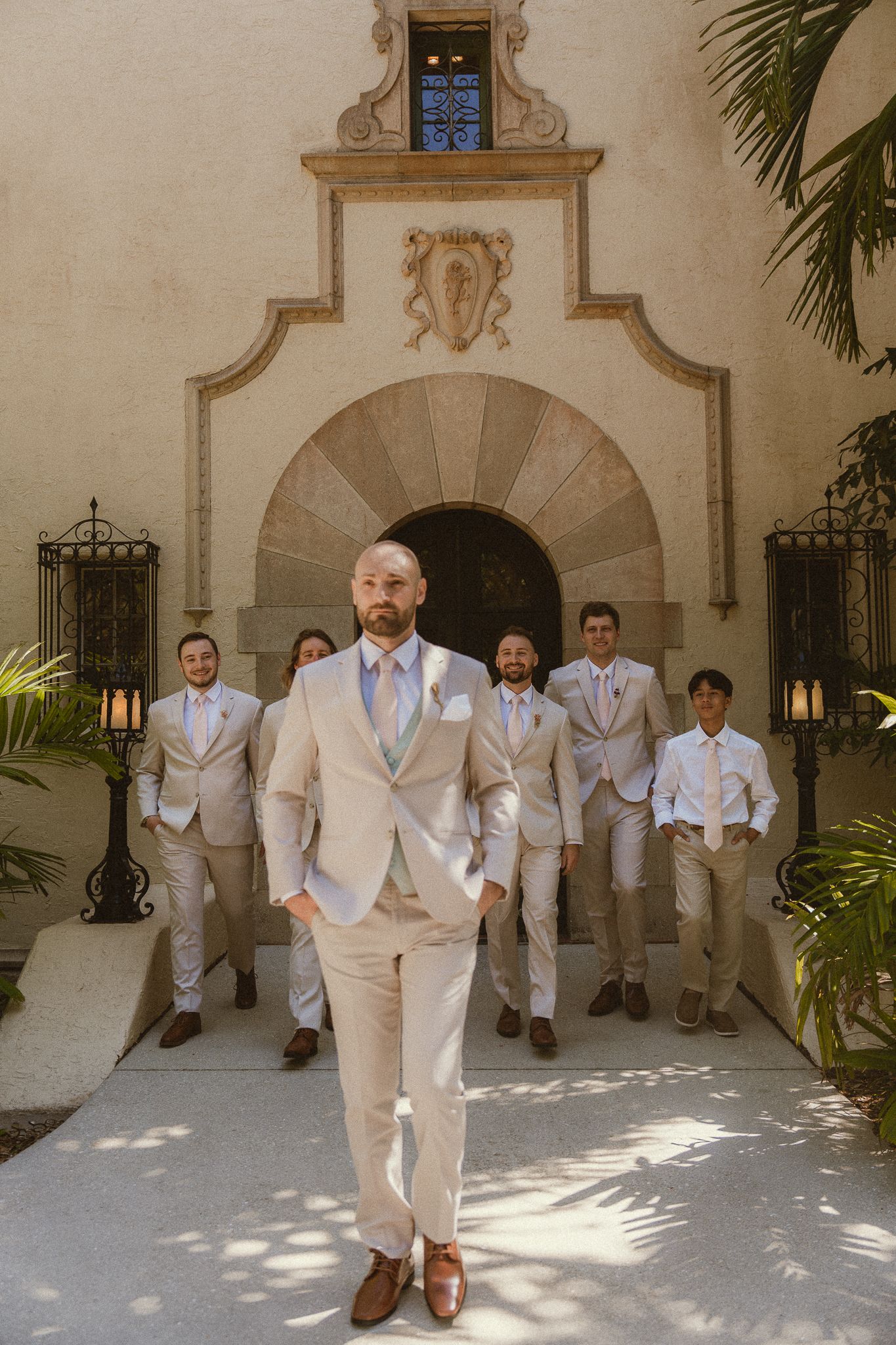 Groom in beige suit leads groomsmen outside elegant Spanish-style venue with ornate archway and iron lanterns.
