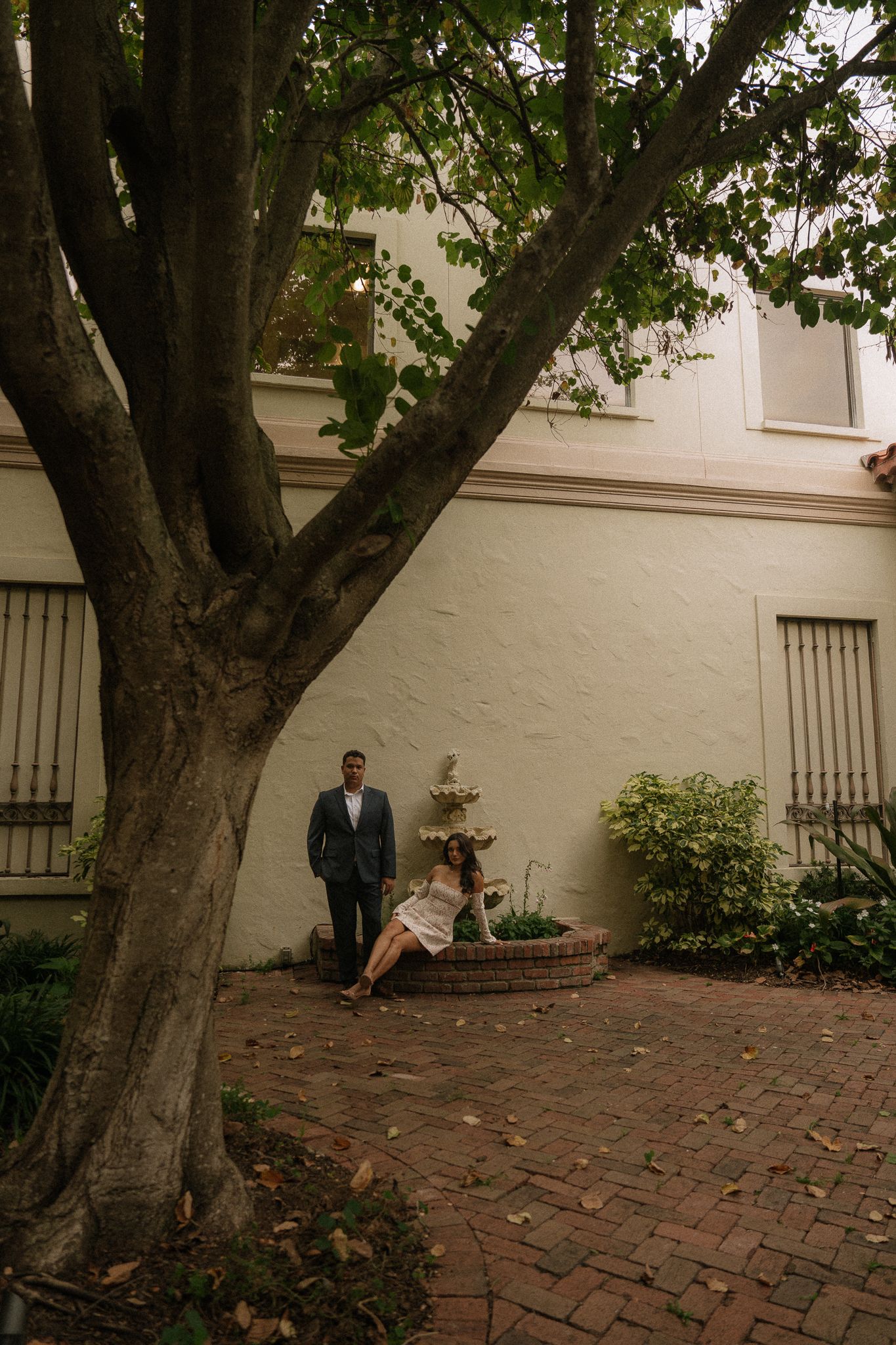 Couple poses near a large tree in a brick courtyard beside a white building with lush greenery.
