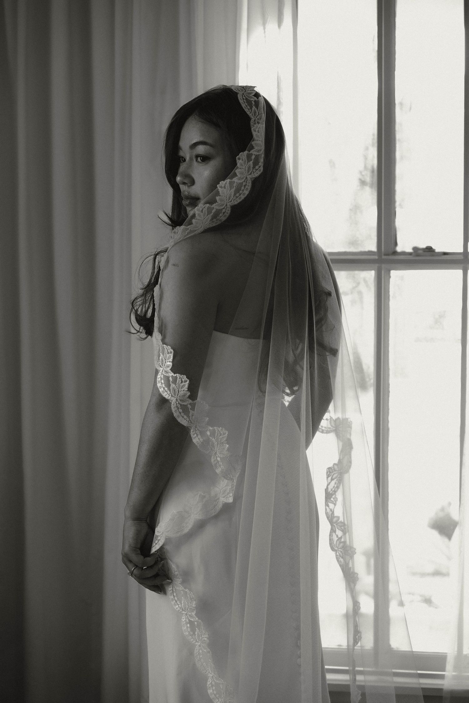 Bride in lace-trimmed veil and wedding gown poses elegantly by window in black and white portrait.