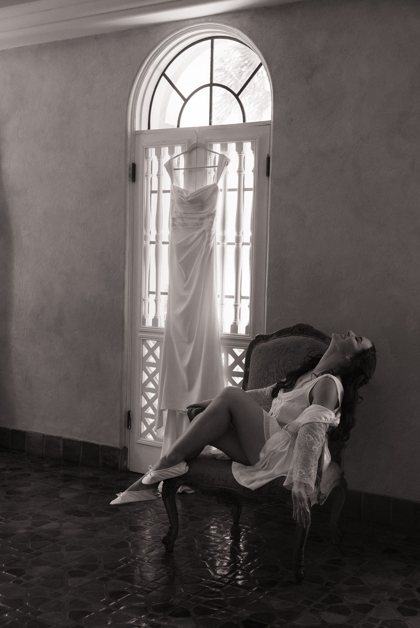 Black and white photo of a bride in lingerie relaxing on a chair with her wedding dress hanging in the background.