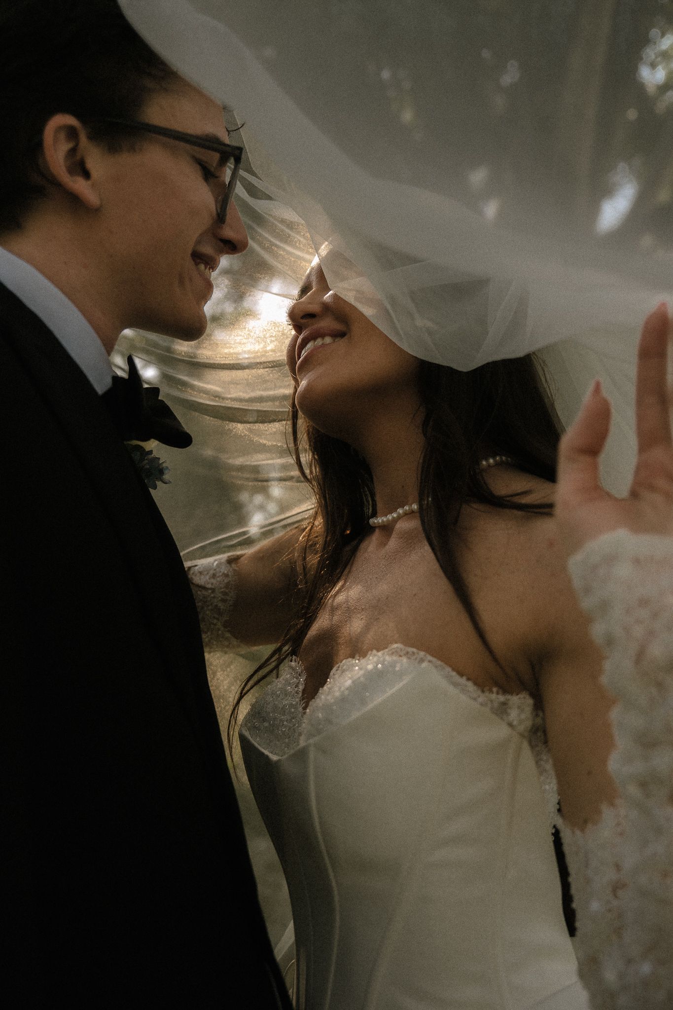 Joyful bride and groom share an intimate moment beneath flowing veil in warm dappled garden light.