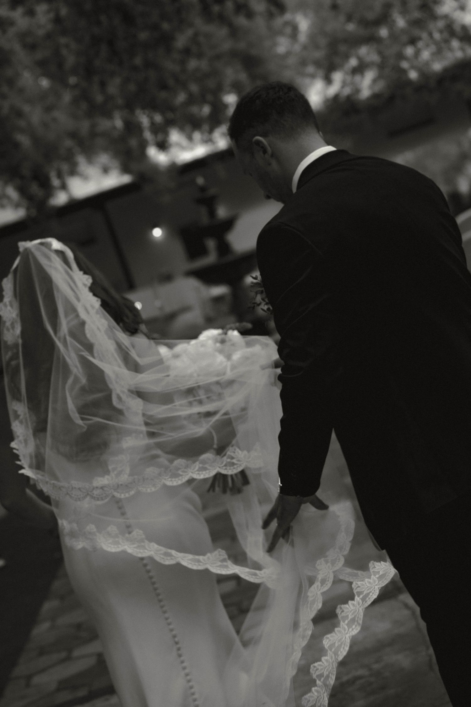 Black and white photo of bride and groom embracing outdoors, veil flowing gracefully in the breeze.