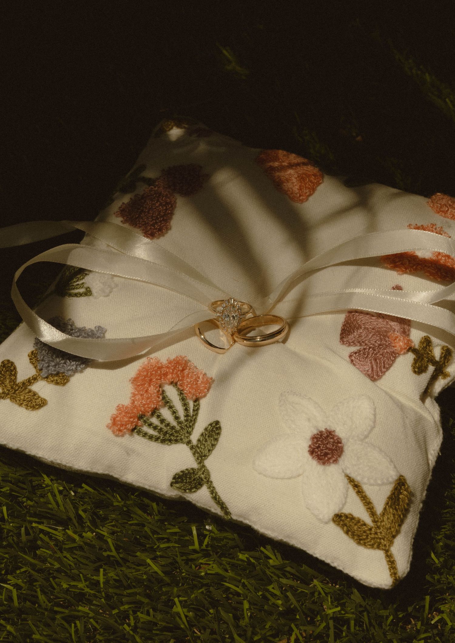 Wedding rings rest on embroidered floral ring bearer pillow with ribbon ties on green grass.